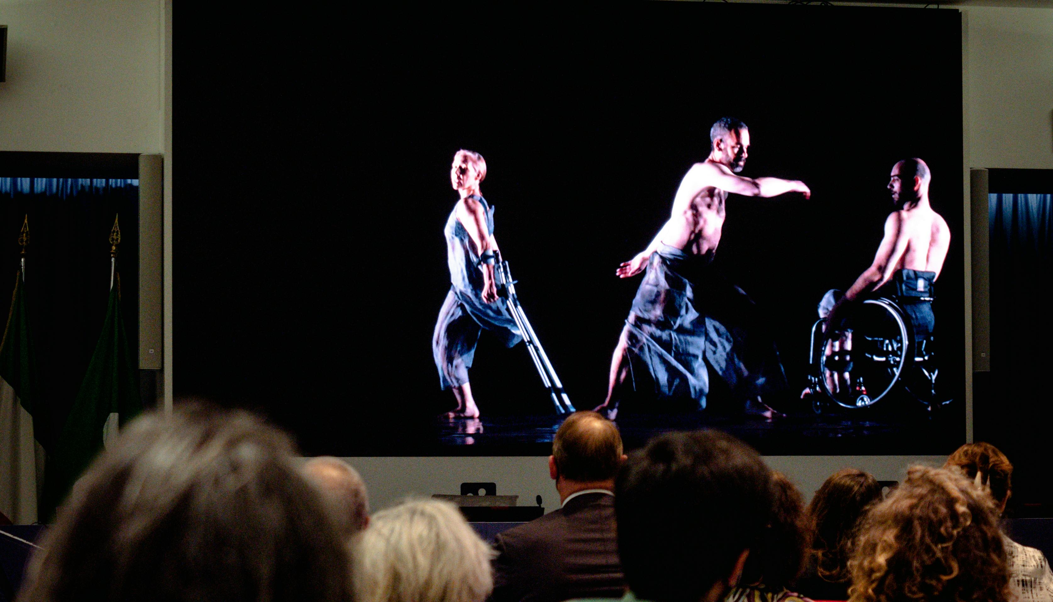 Pubblico guarda un video in proiezione con una danzatrice e due danzatori con disabilità.
