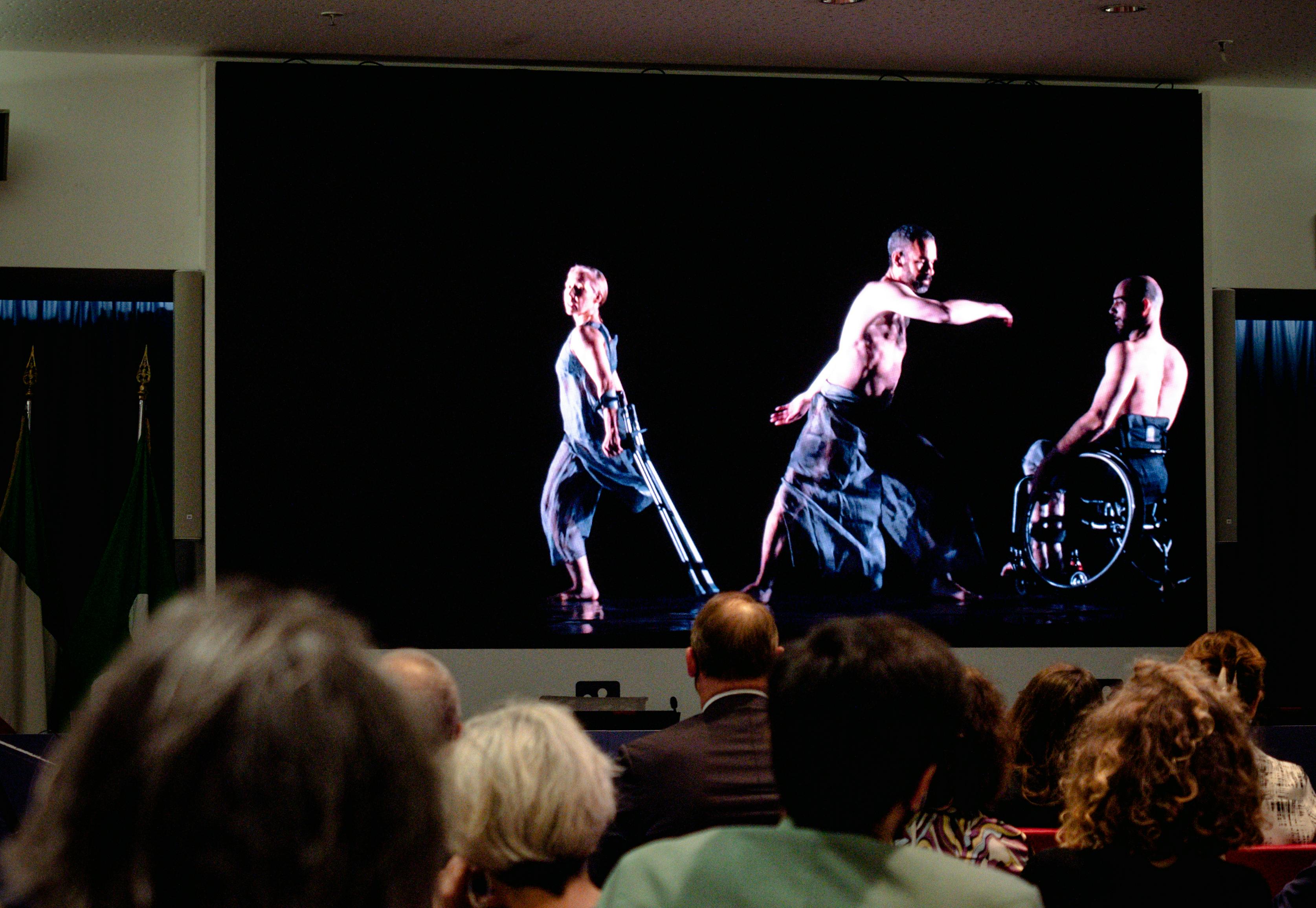 Pubblico guarda un video in proiezione con una danzatrice e due danzatori con disabilità.