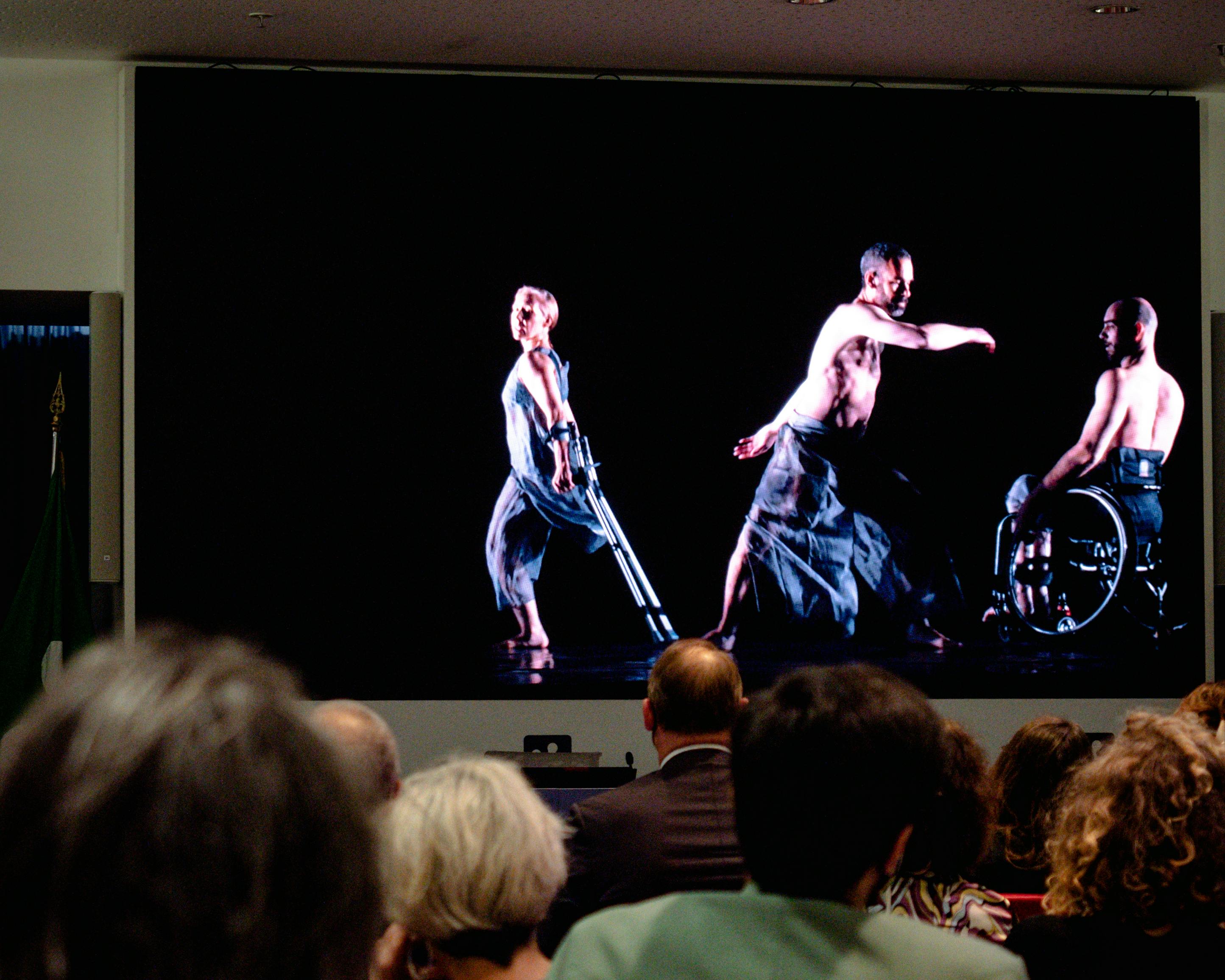 Pubblico guarda un video in proiezione con una danzatrice e due danzatori con disabilità.