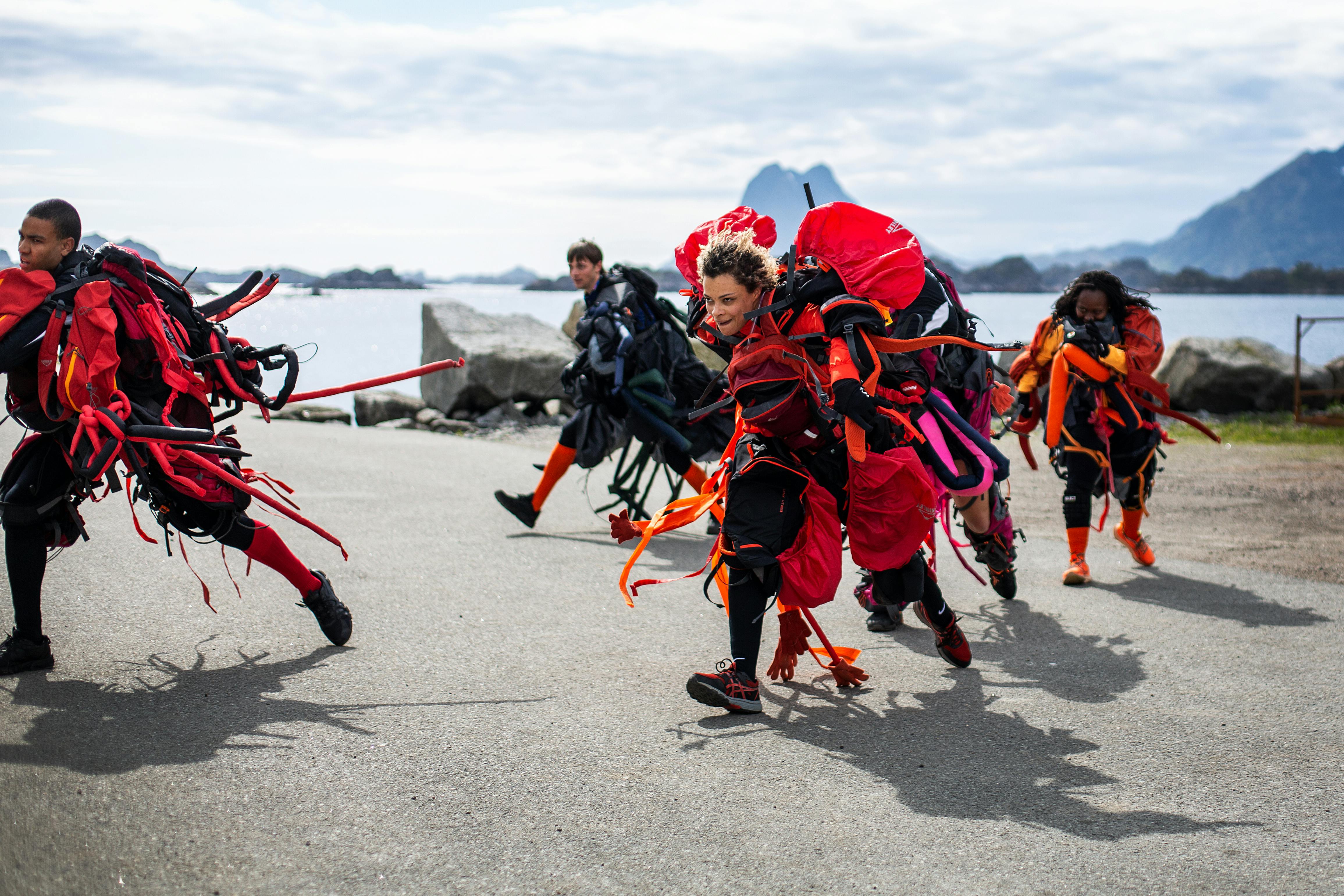 Five male and female dancers in eye-catching costumes, in an open setting with mountains in the background.