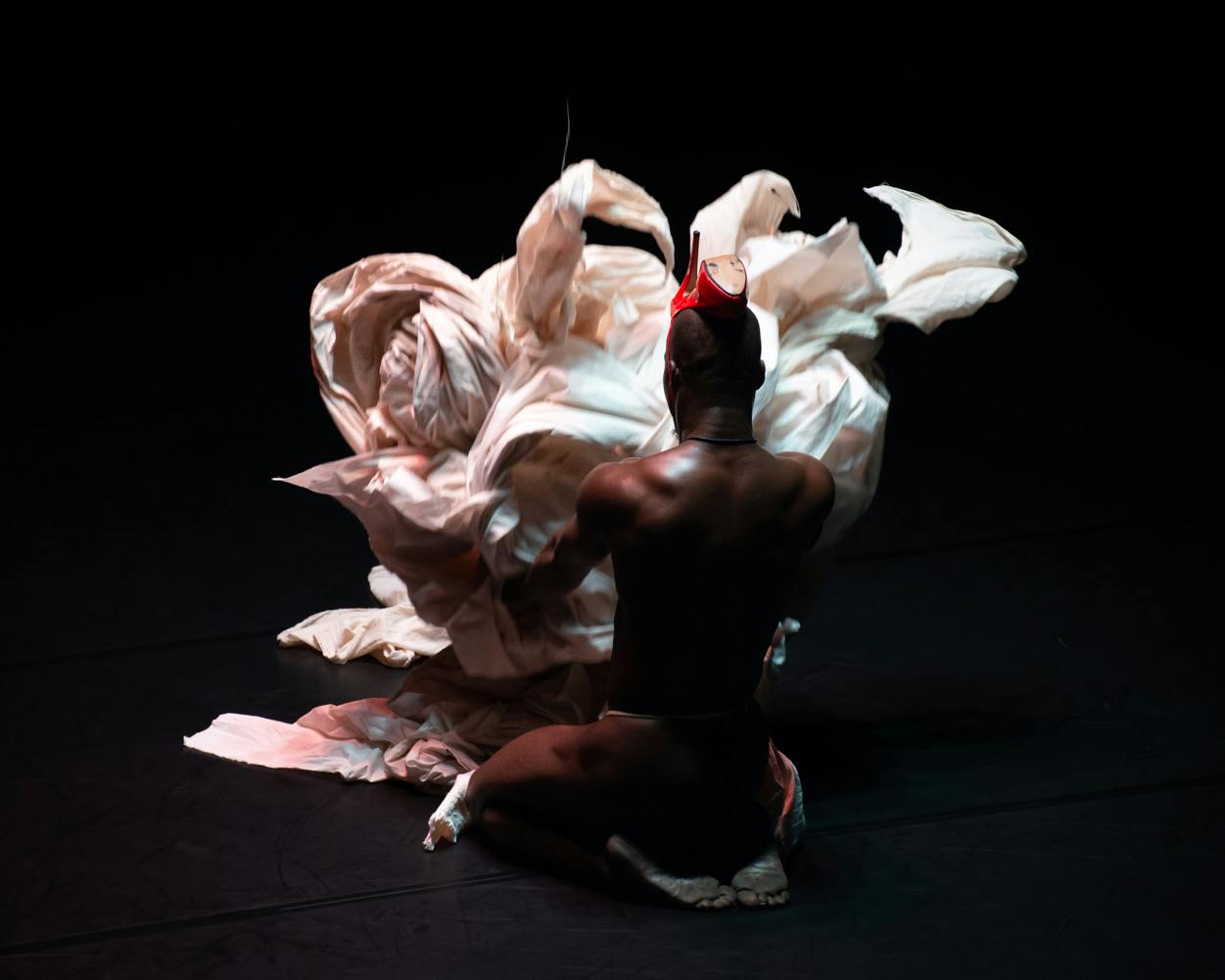 Un ballerino a dorso nudo è inginocchiato di spalle e gioca con delle lenzuola bianche.