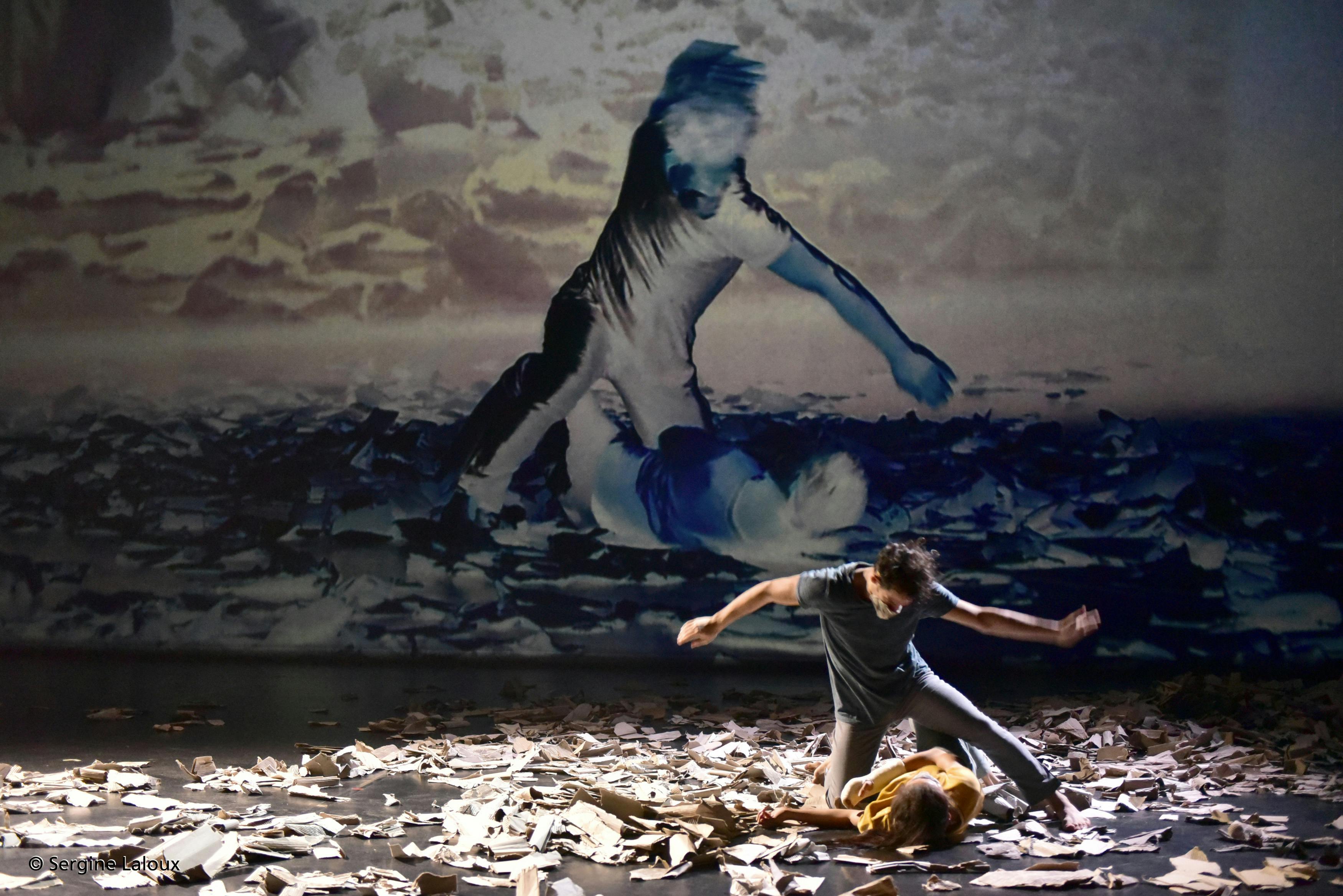 A dancer lying on the stage with sheets of paper and a dancer kneeling beside her. In the background the same image in video projection.