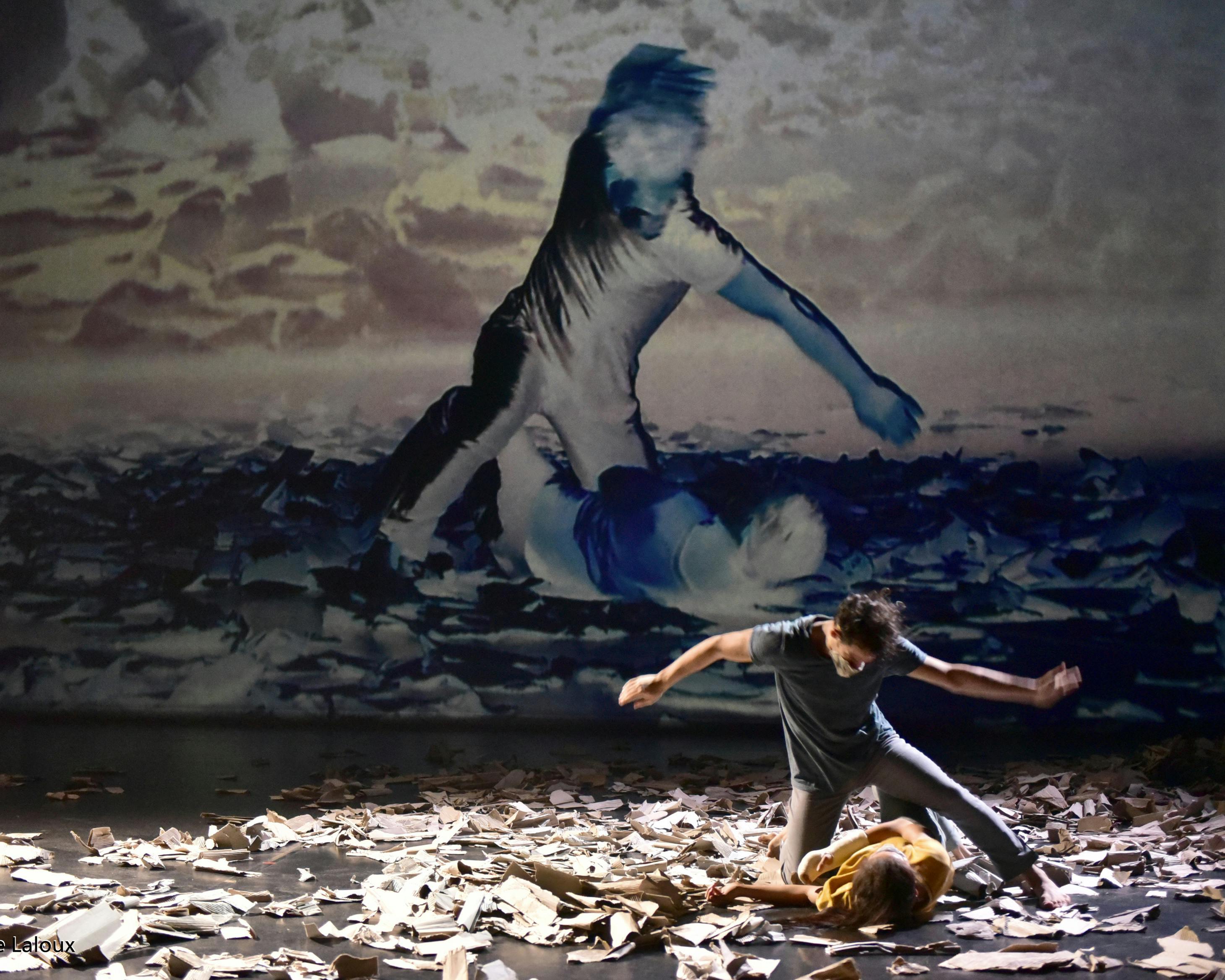 A dancer lying on the stage with sheets of paper and a dancer kneeling beside her. In the background the same image in video projection.