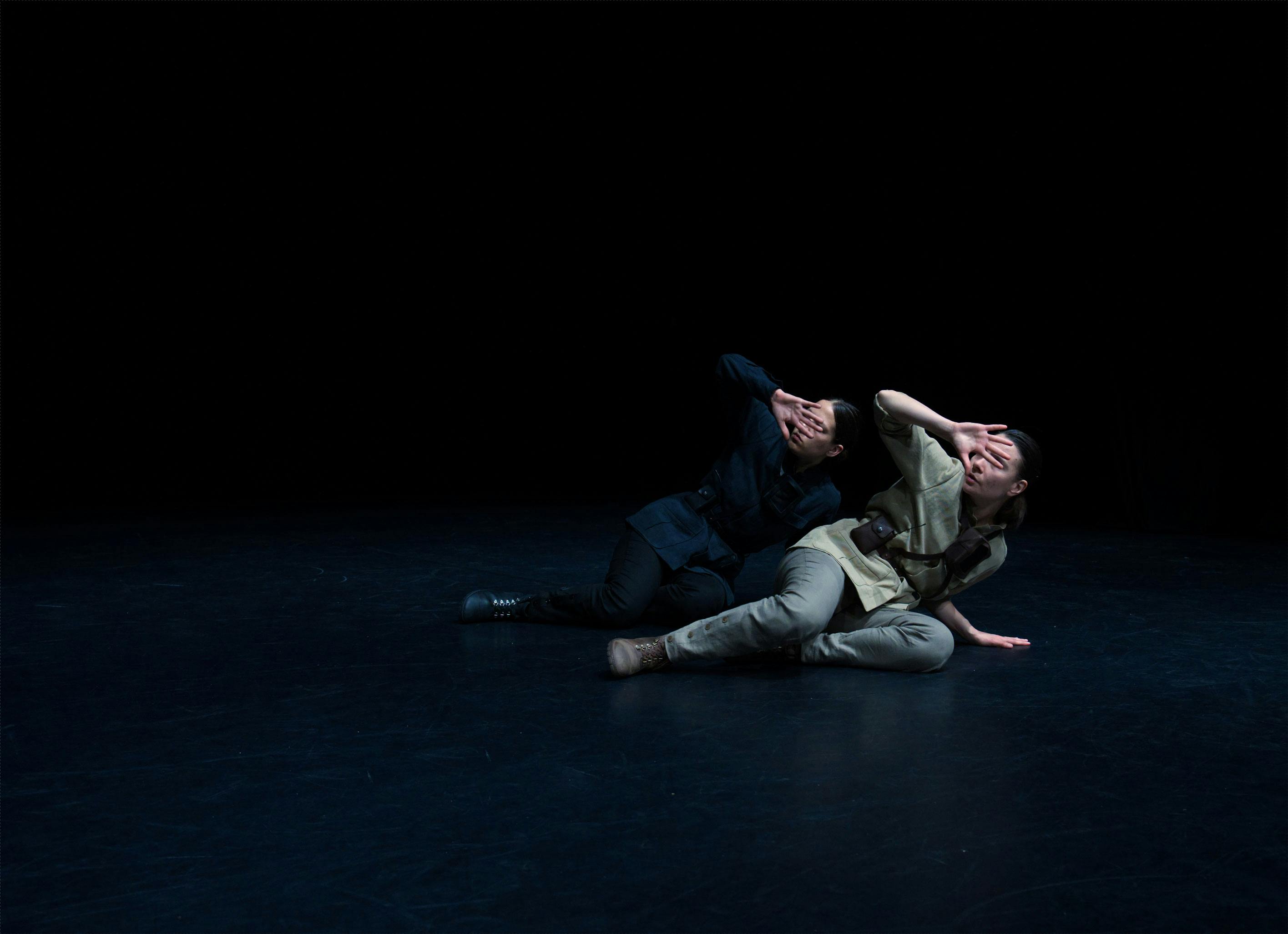 Two dancers semi-reclining in front of each other, cover their faces with one hand as if protecting themselves from a light.