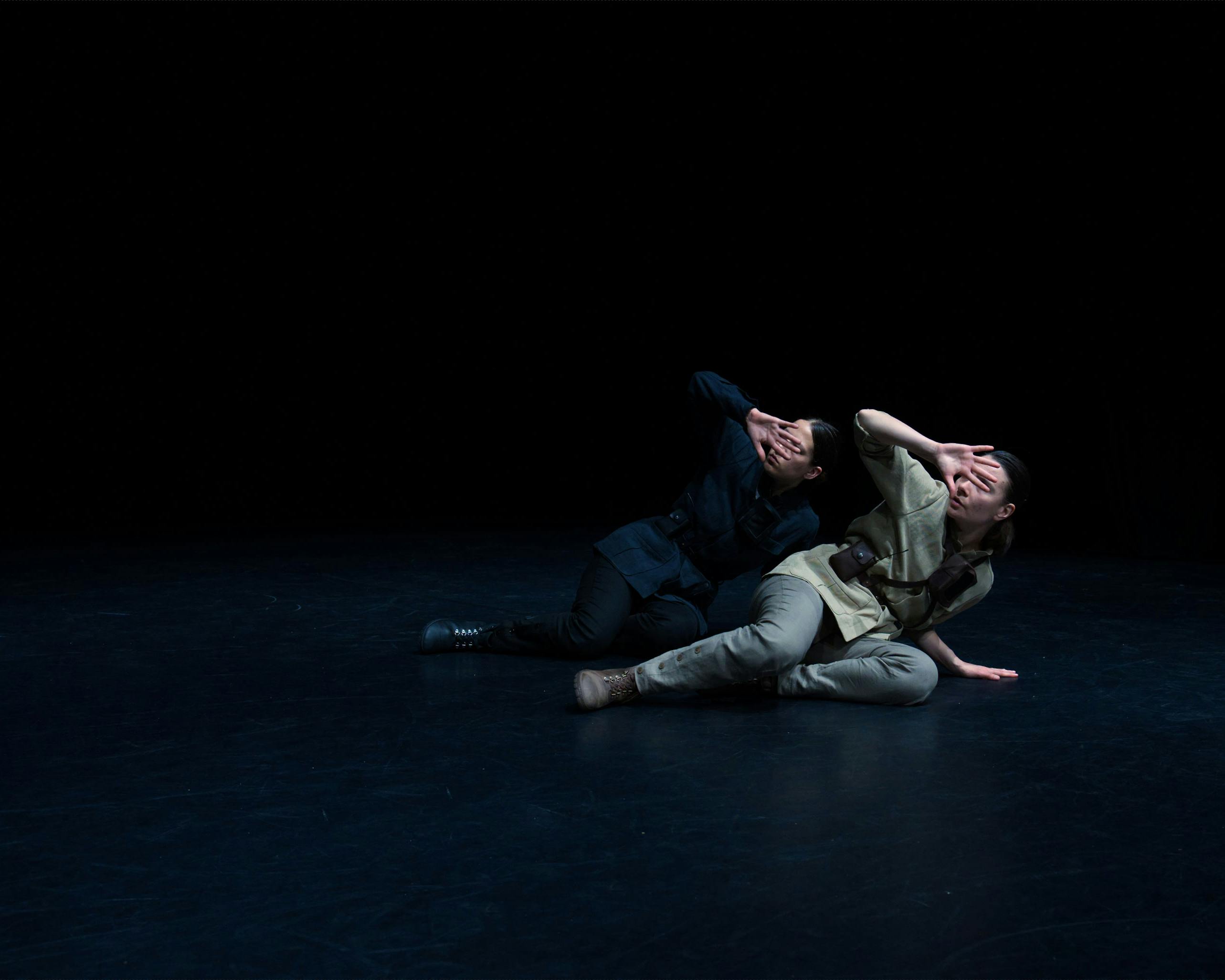Two dancers semi-reclining in front of each other, cover their faces with one hand as if protecting themselves from a light.