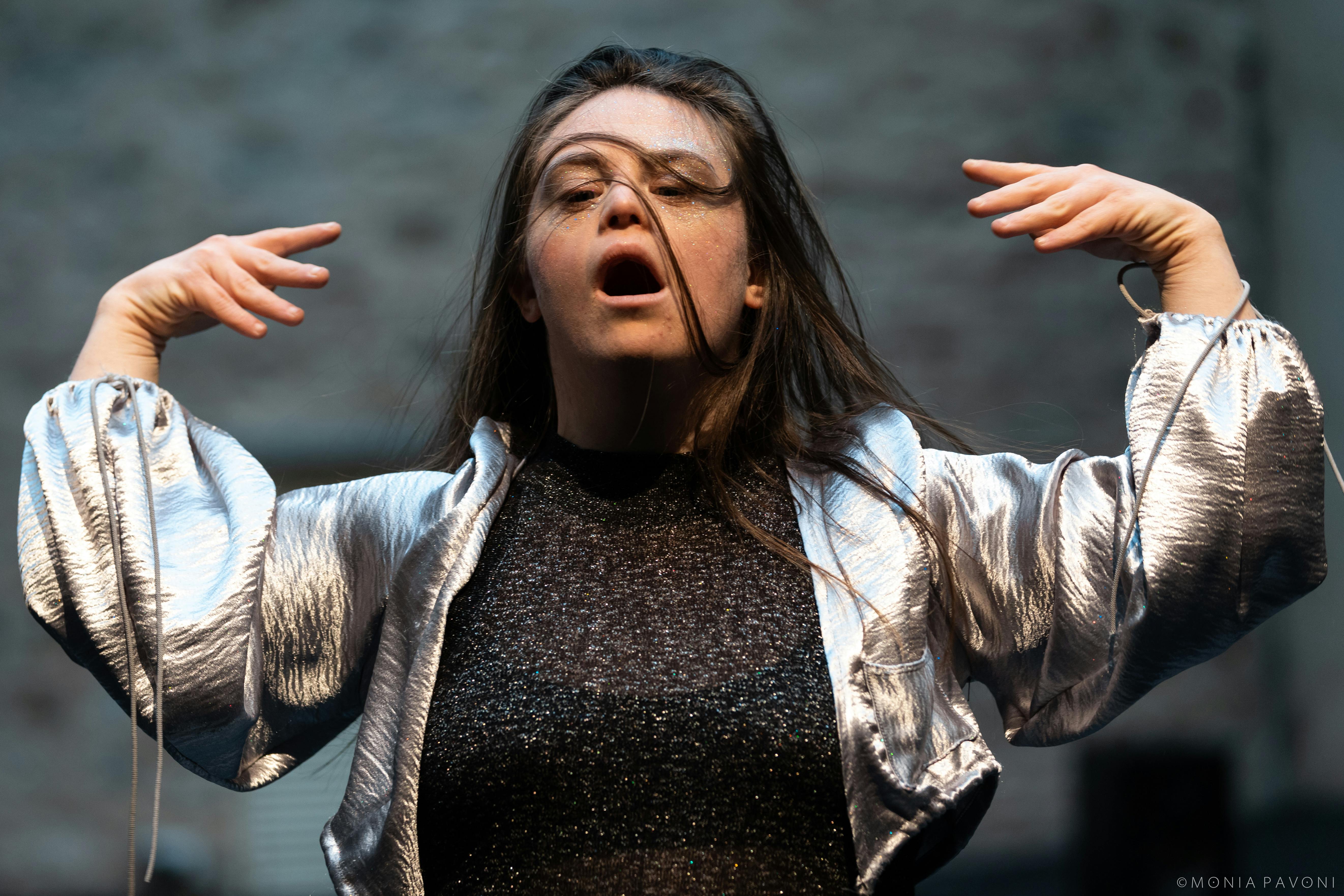 The performer with disabilities has her arms out to the side with her hands pointing towards her head, wears a silver jacket