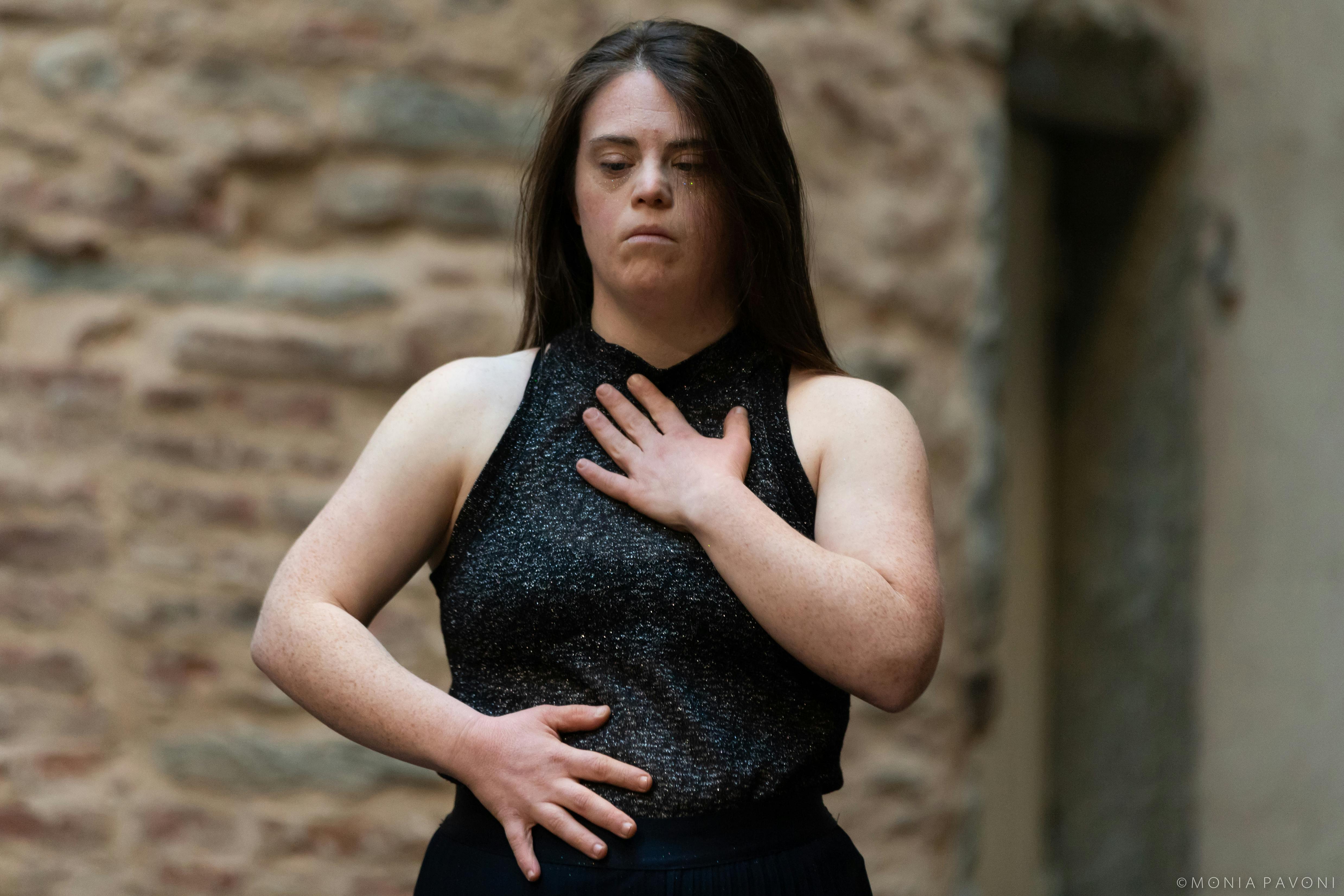 la performer con disabilità ha una mano sul petto e una sul fianco indossa una canottiera grigia