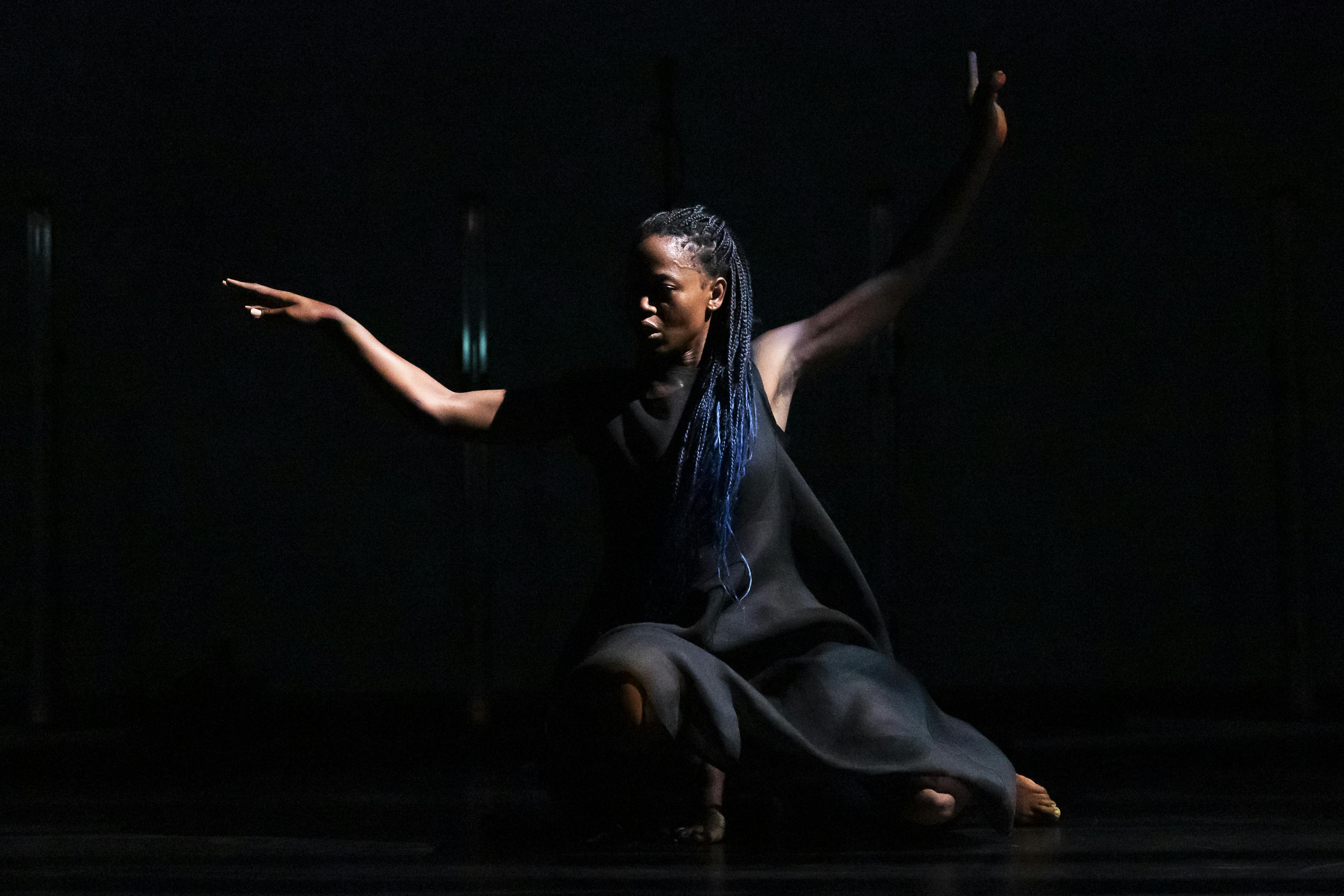 Una danzatrice vestita di nero è quasi inginocchiata con entrambe le braccia che cercano l'equilibrio.
