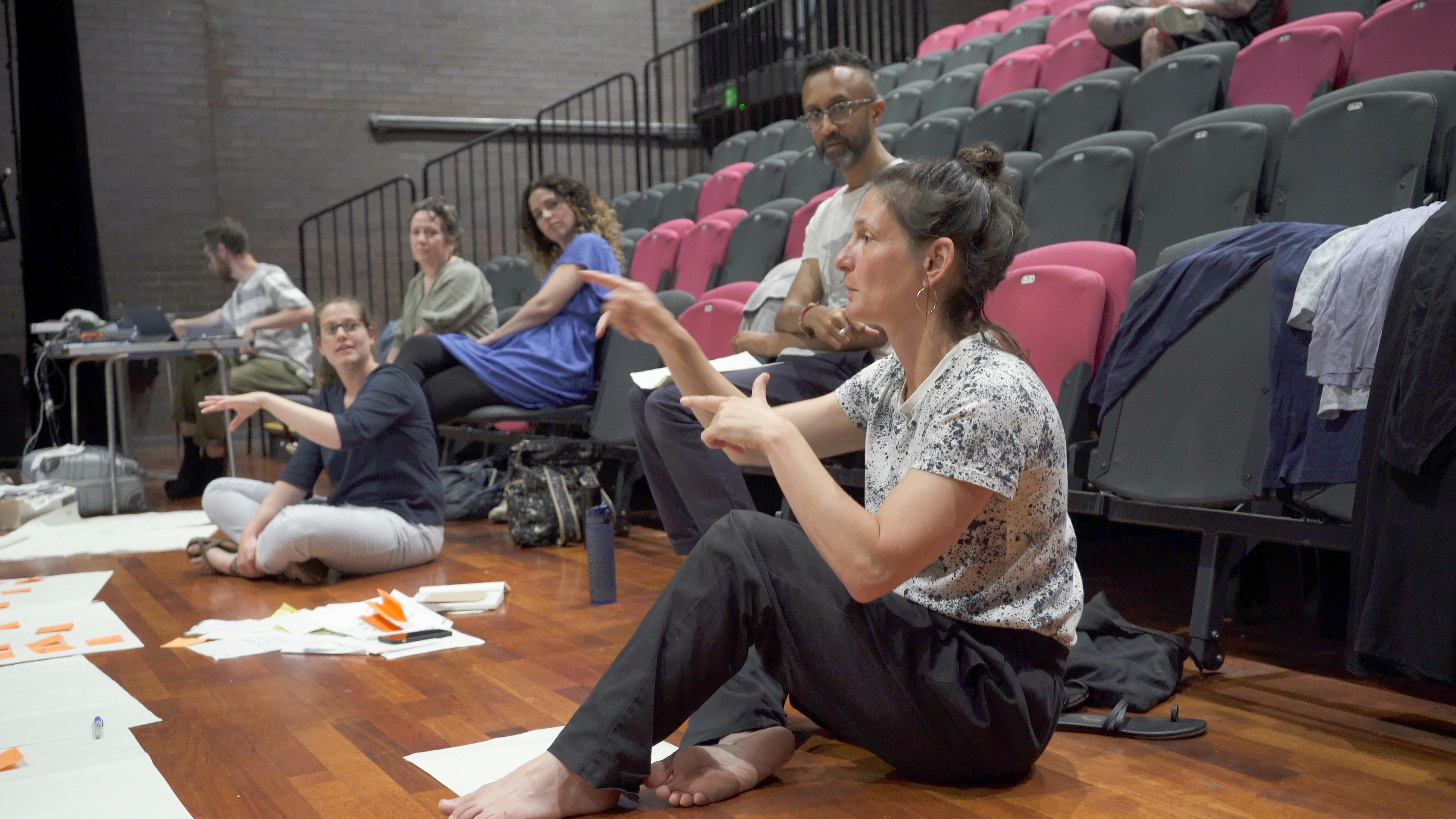 The artist Louise Stern sitting on the floor, in the background five people observe her