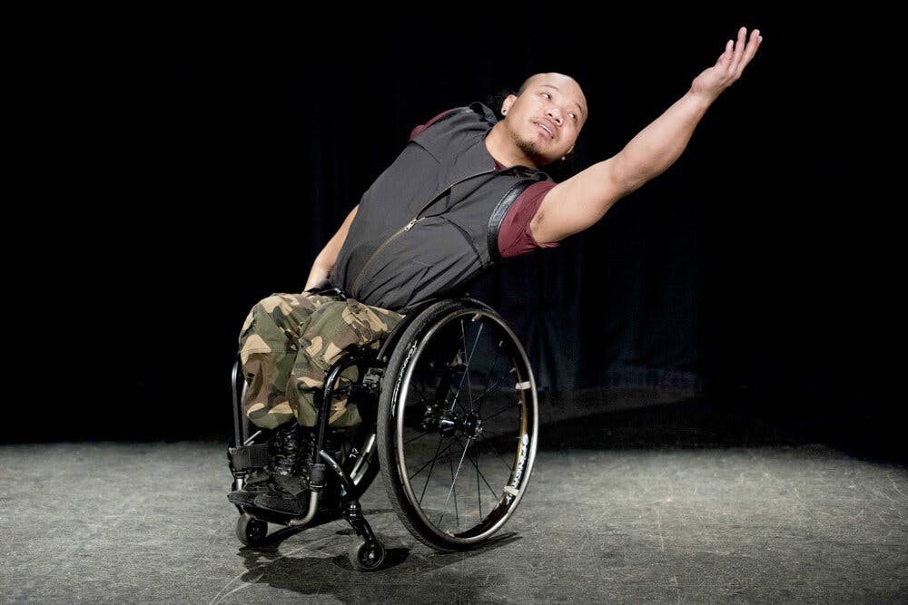 The wheelchair-bound artist Nadenh Poan stands on stage with her arm extended to her left