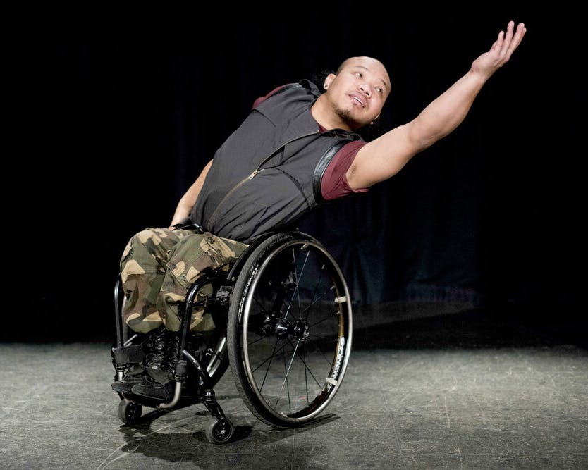 The wheelchair-bound artist Nadenh Poan stands on stage with her arm extended to her left