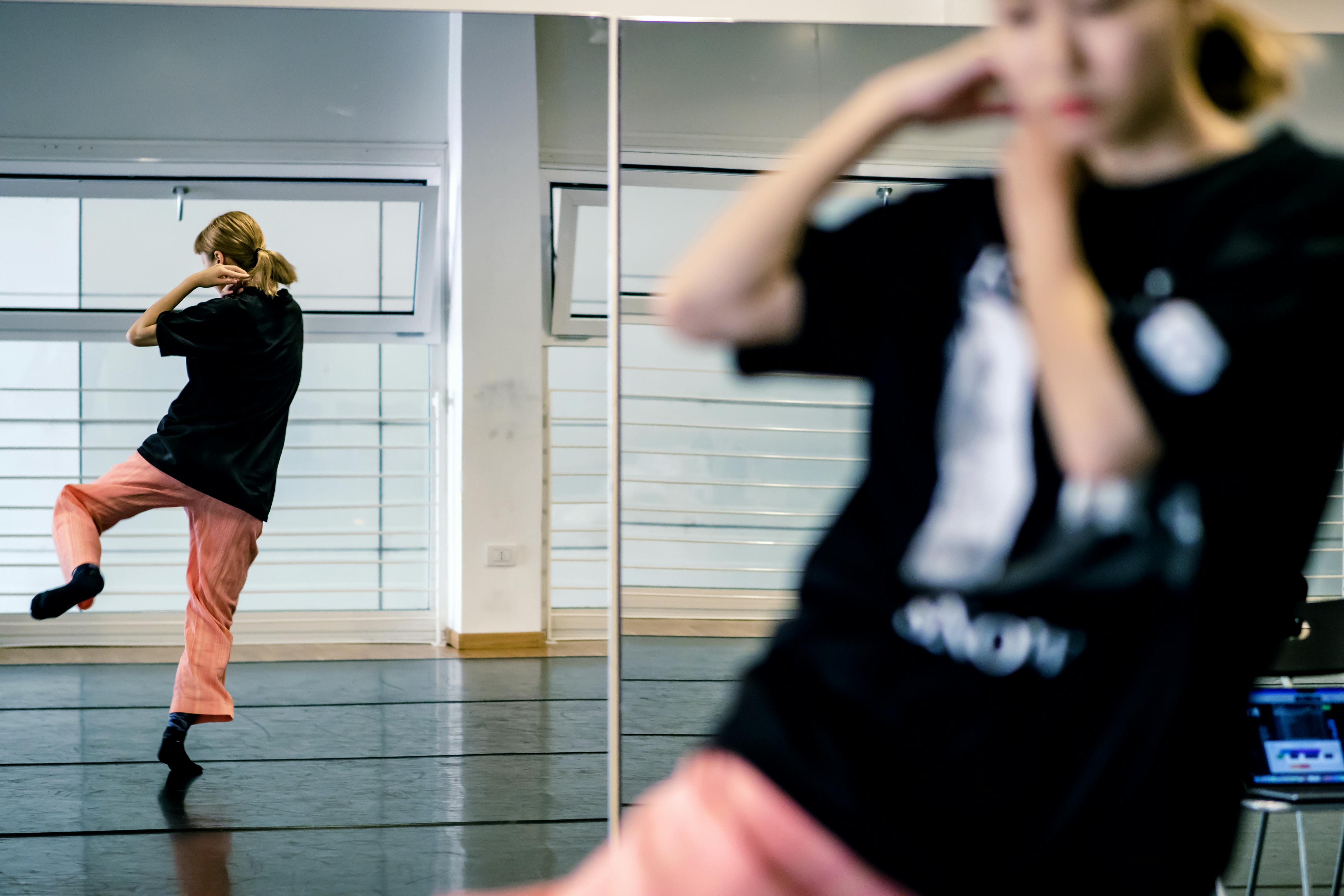 The dancer Yoko Omori in pink trousers and a black T-shirt in the foreground is out of focus. Her back is reflected in a mirror. She dances with one leg raised while both hands approach one shoulder