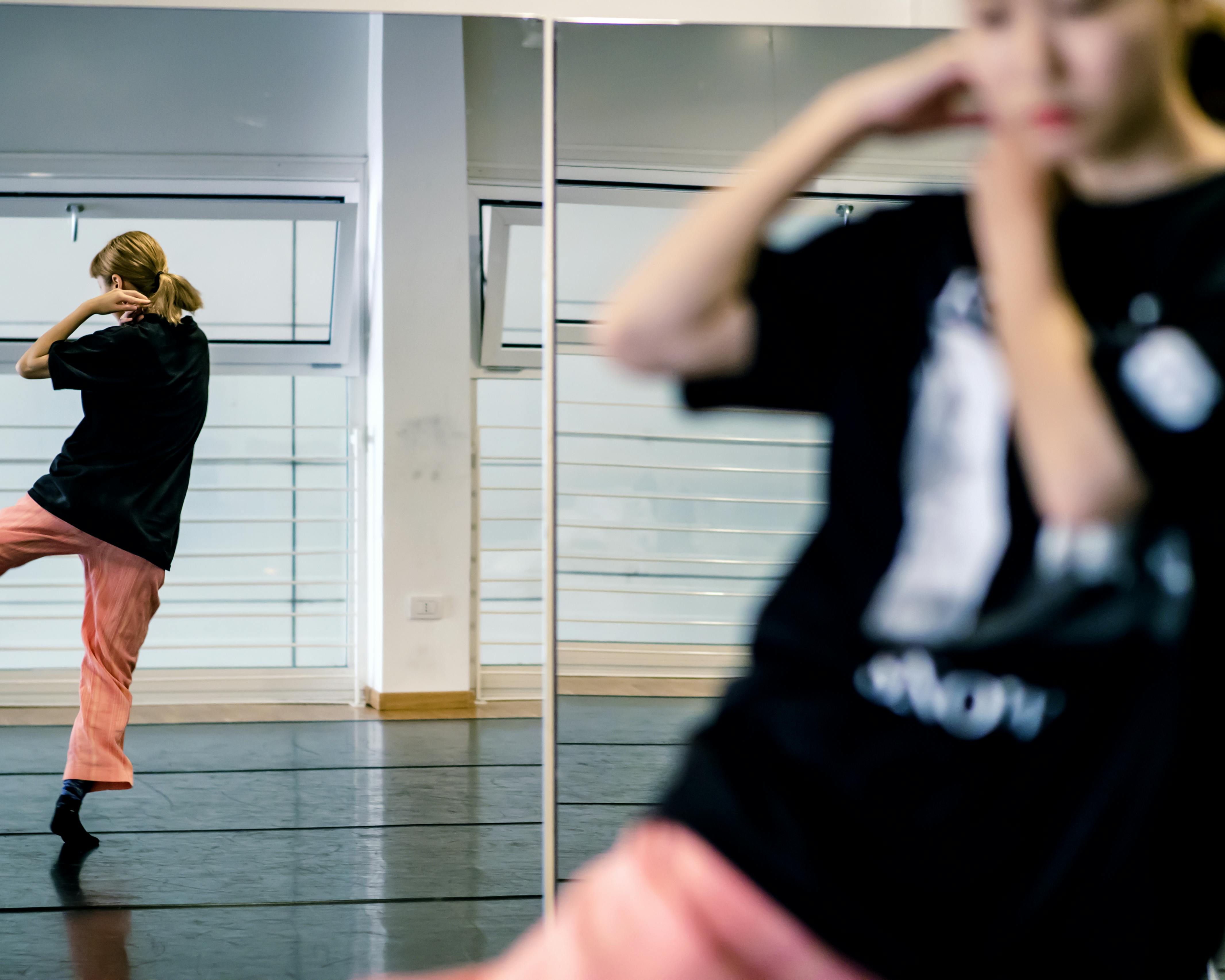The dancer Yoko Omori in pink trousers and a black T-shirt in the foreground is out of focus. Her back is reflected in a mirror. She dances with one leg raised while both hands approach one shoulder