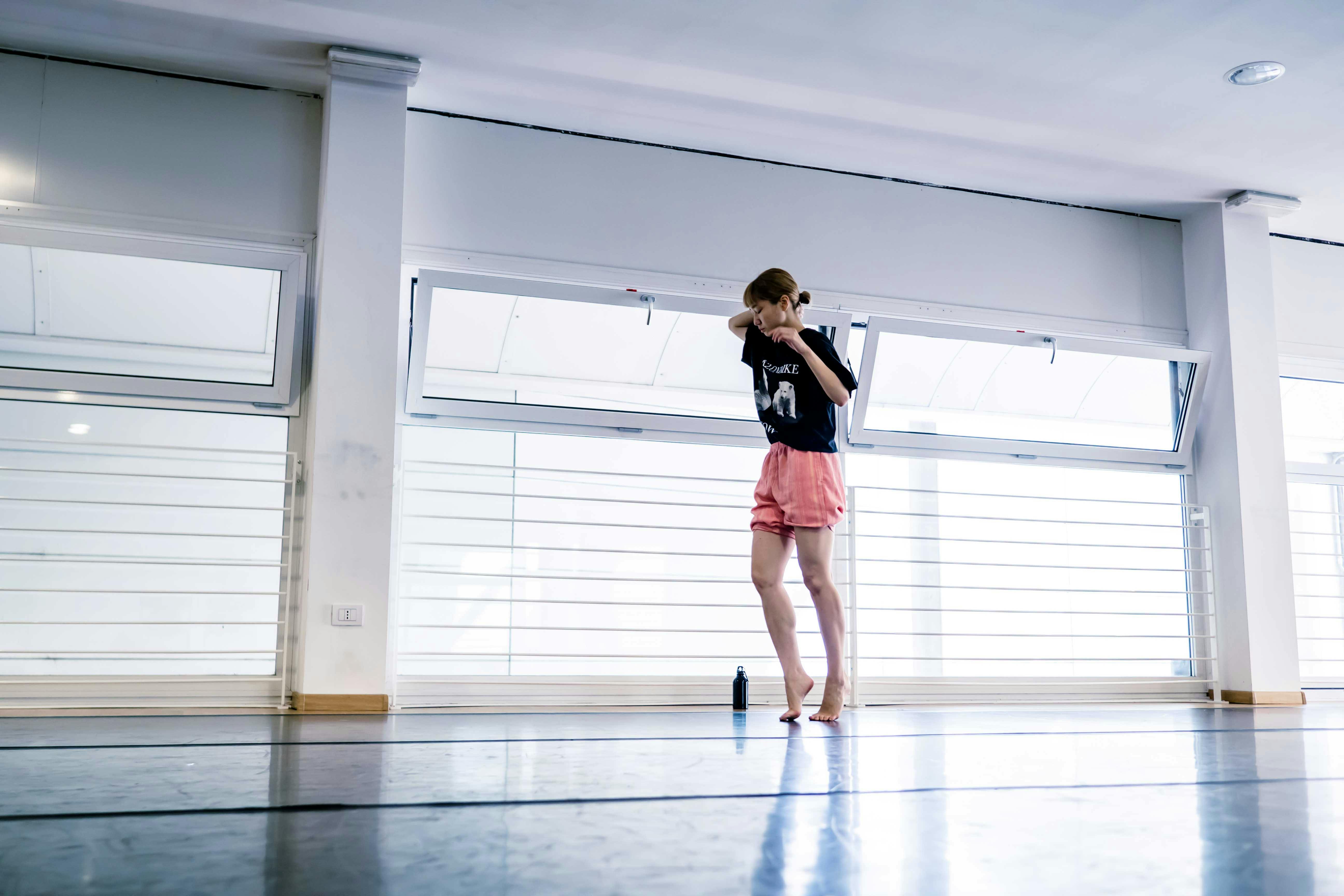 La danzatrice Yoko Omori durante le prove in sala, mentre cammina in punta di piedi con i gomiti sollevati e la mani vicino al viso.