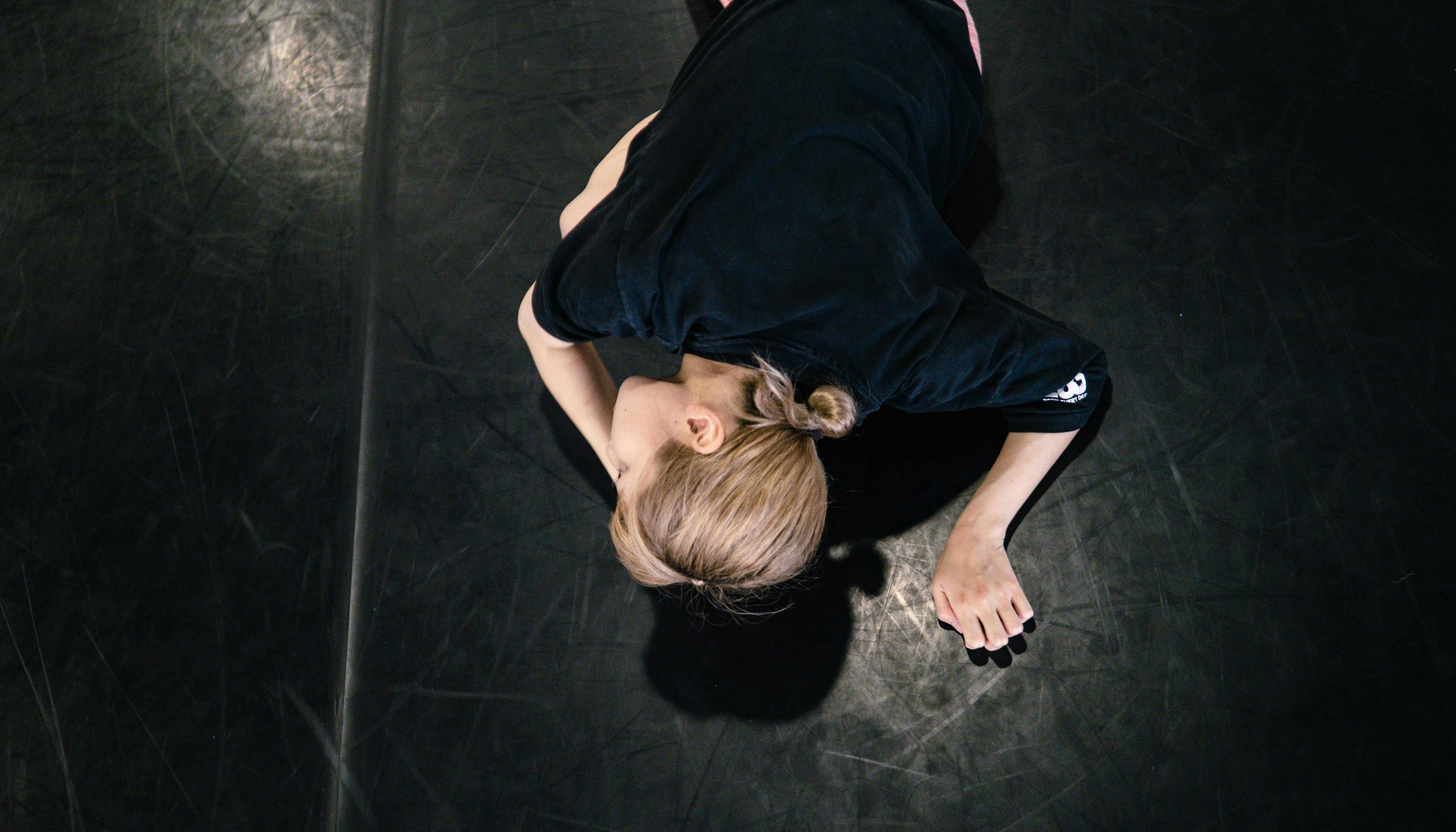La danzatrice Yoko Omori ripresa dall'alto mentre è sdraiata sul pavimento a pancia in giù con le mani vicino alla testa.
