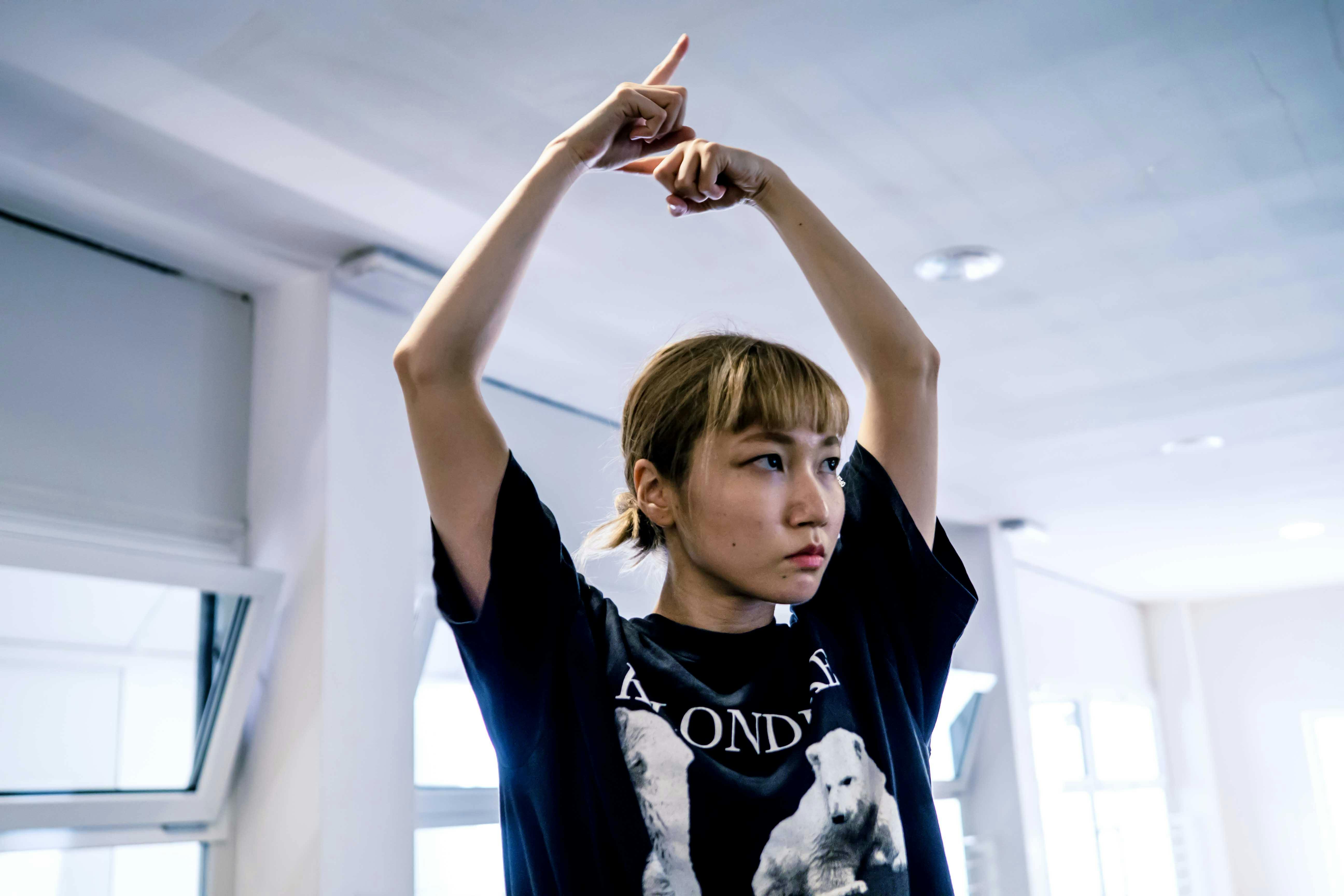 La danzatrice Yoko Omori ripresa a mezzo busto, con lo sguardo rivolto altrove ed entrambe le braccia sollevate e le mani riprese in un movimento circolare con l'indice puntato.