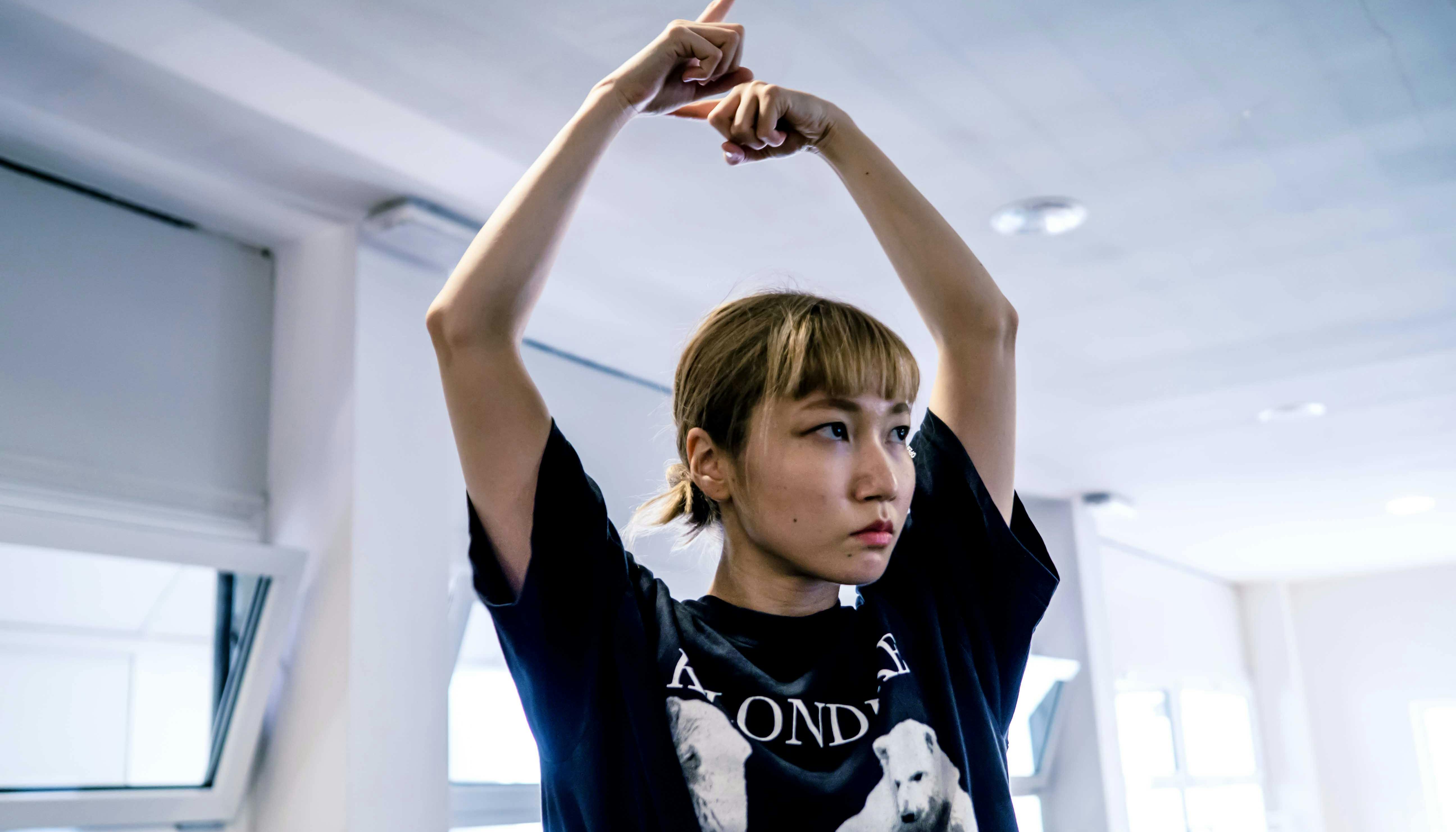 La danzatrice Yoko Omori ripresa a mezzo busto, con lo sguardo rivolto altrove ed entrambe le braccia sollevate e le mani riprese in un movimento circolare con l'indice puntato.