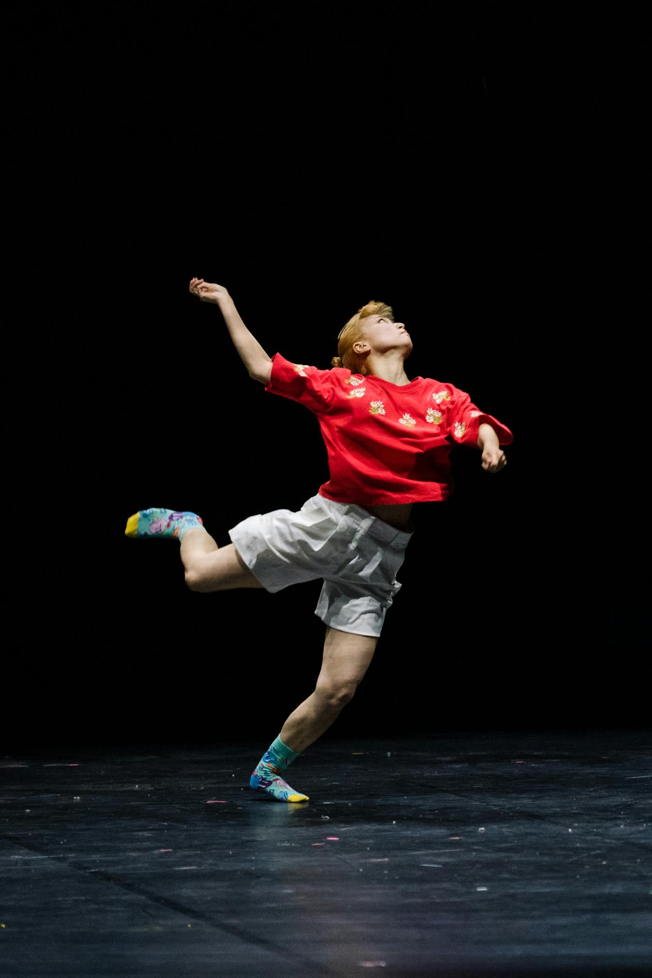 Performer in a red shirt and colorful socks, has the weight only on his left leg and his right arm raised.