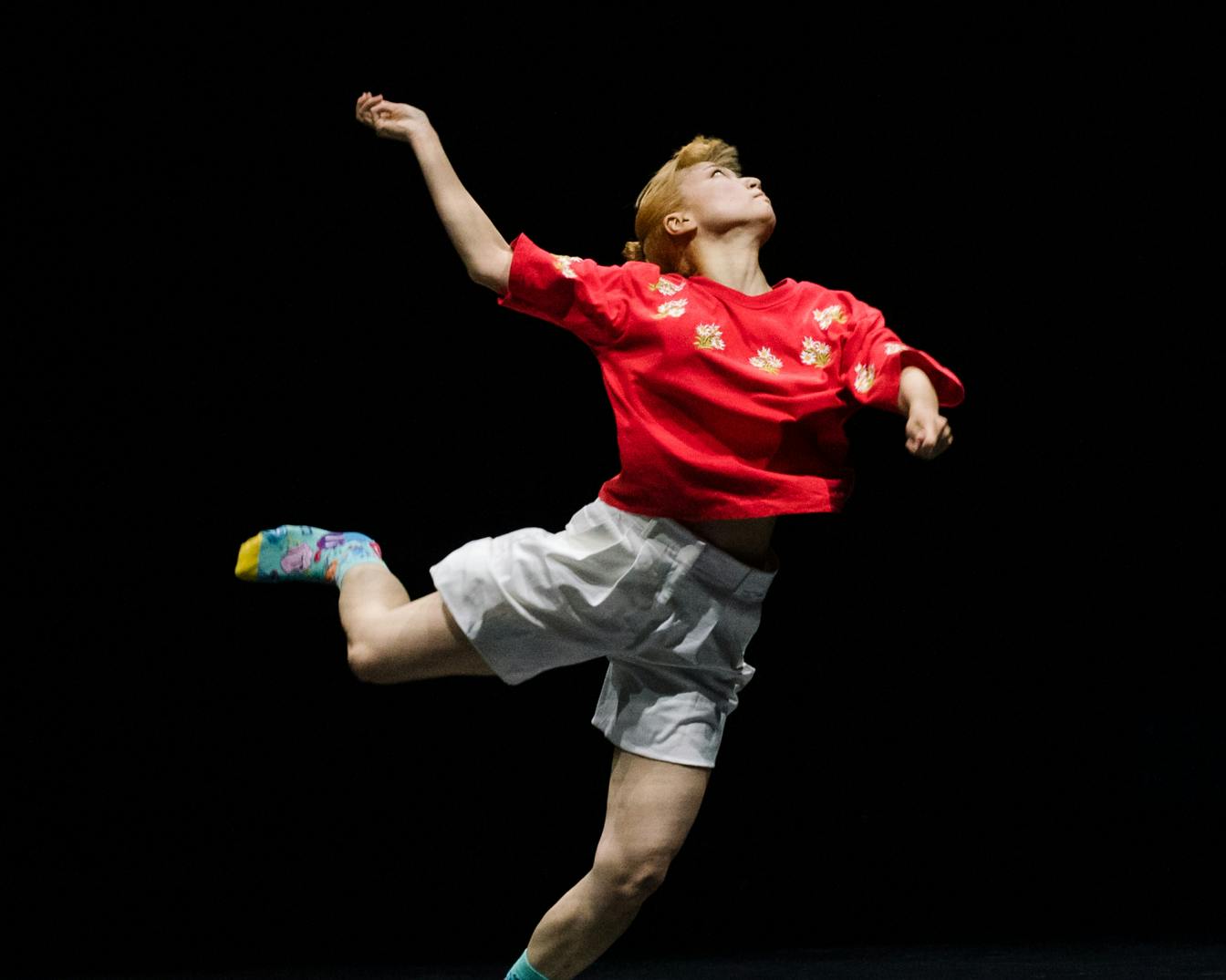 Performer in a red shirt and colorful socks, has the weight only on his left leg and his right arm raised.
