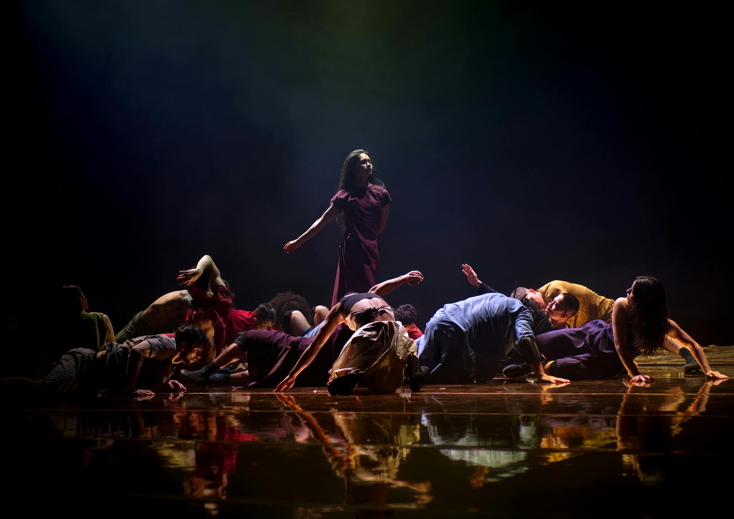 Una danzatrice in piedi con abito viola è circondata da danzatori e danzatrici  inginocchiate o sdraiate