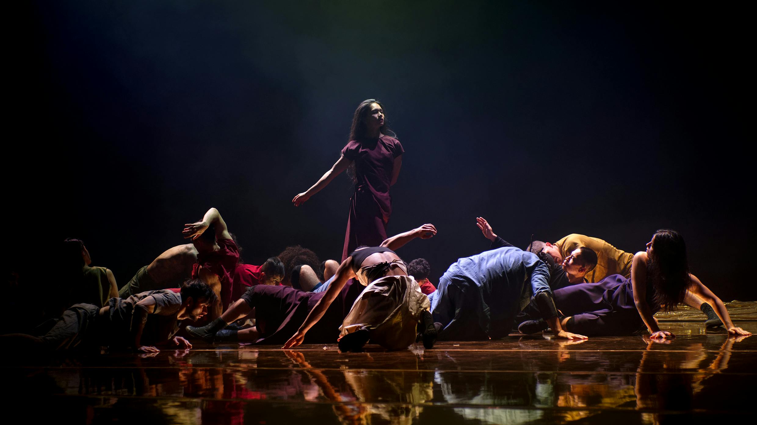 Una danzatrice in piedi con abito viola è circondata da danzatori e danzatrici  inginocchiate o sdraiate