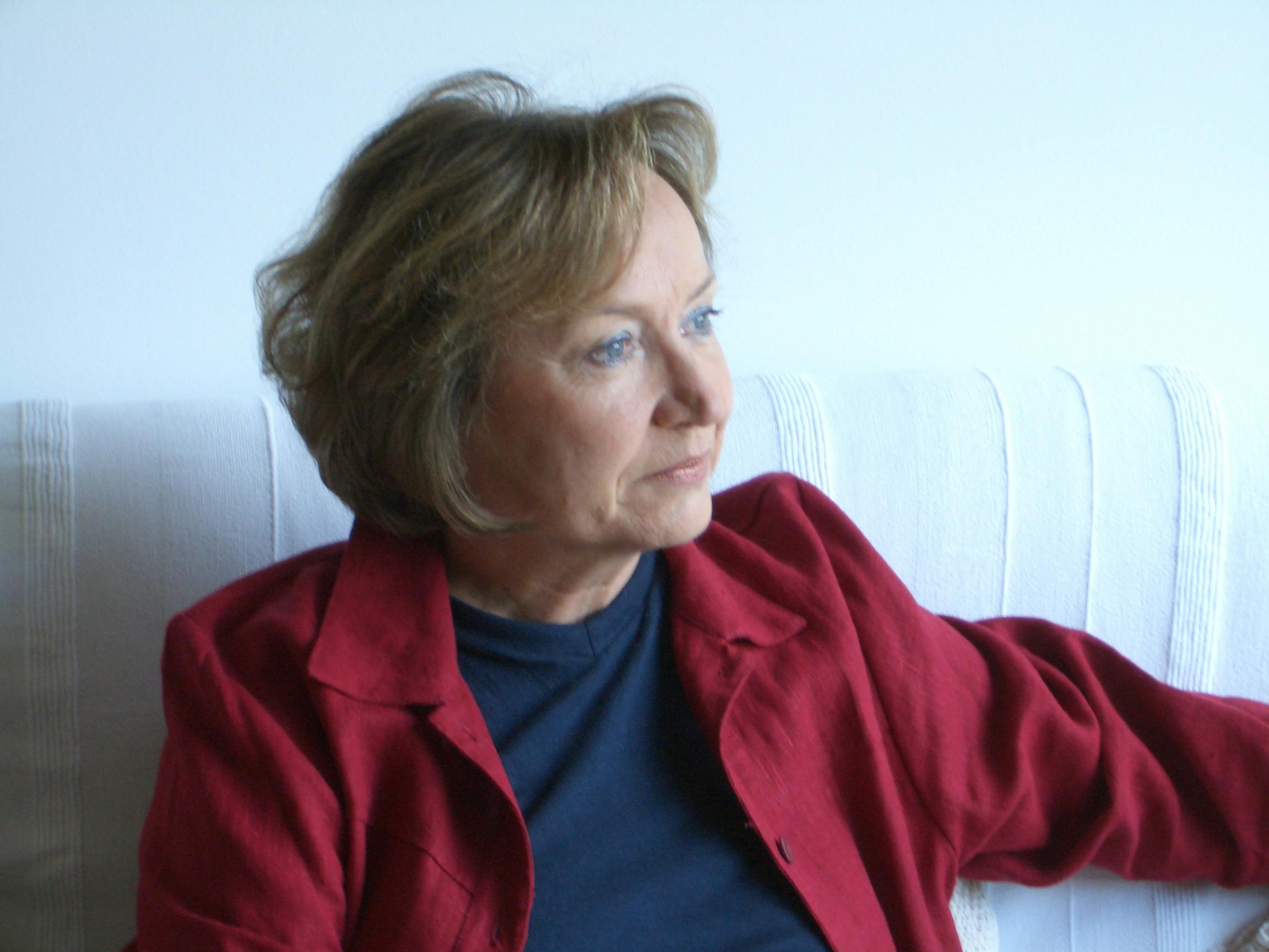 Portrait of Melita Richter. She is looking to her left, sitting on a white sofa. She is wearing a blue sweater and a red shirt.