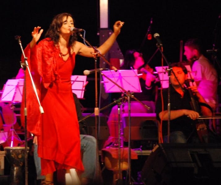 Concert photo. In the foreground, a singer dressed in red. Behind her, seated musicians.