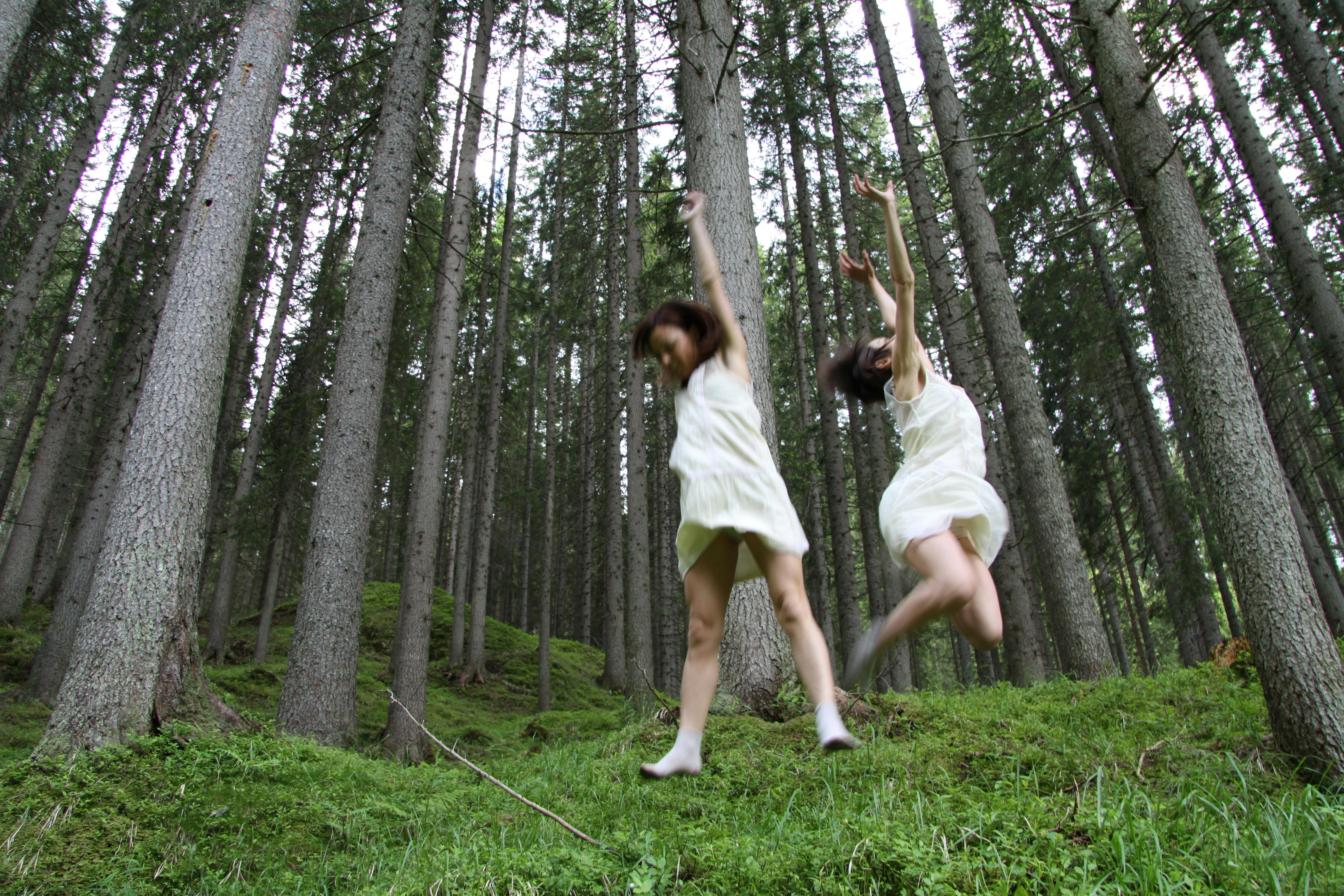 Two people in the woods, surrounded by very tall trees. They are dressed in white and are jumping.