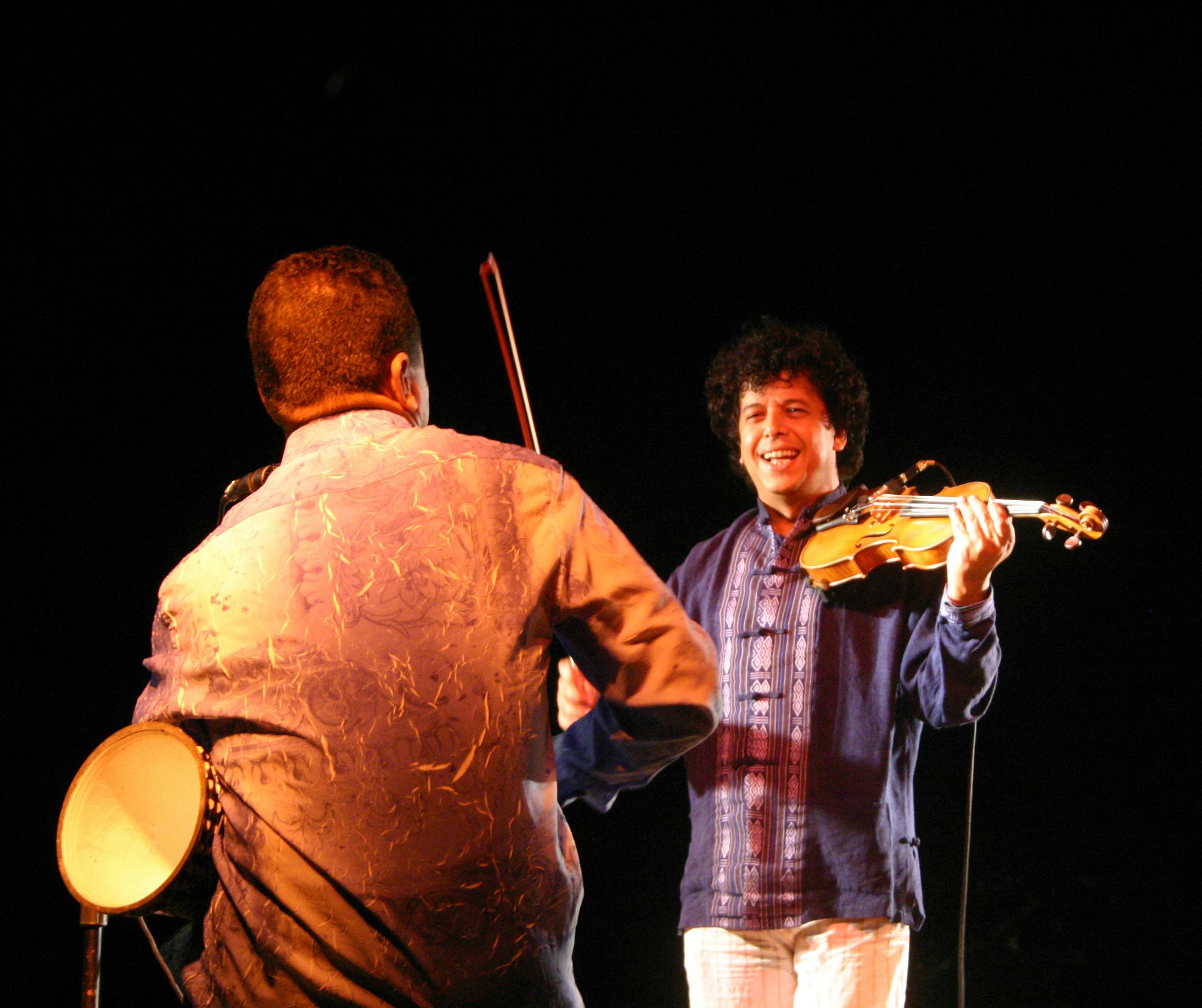 Two musicians. One is facing away, holding a drum under his arm. In front of him, a violinist is smiling.