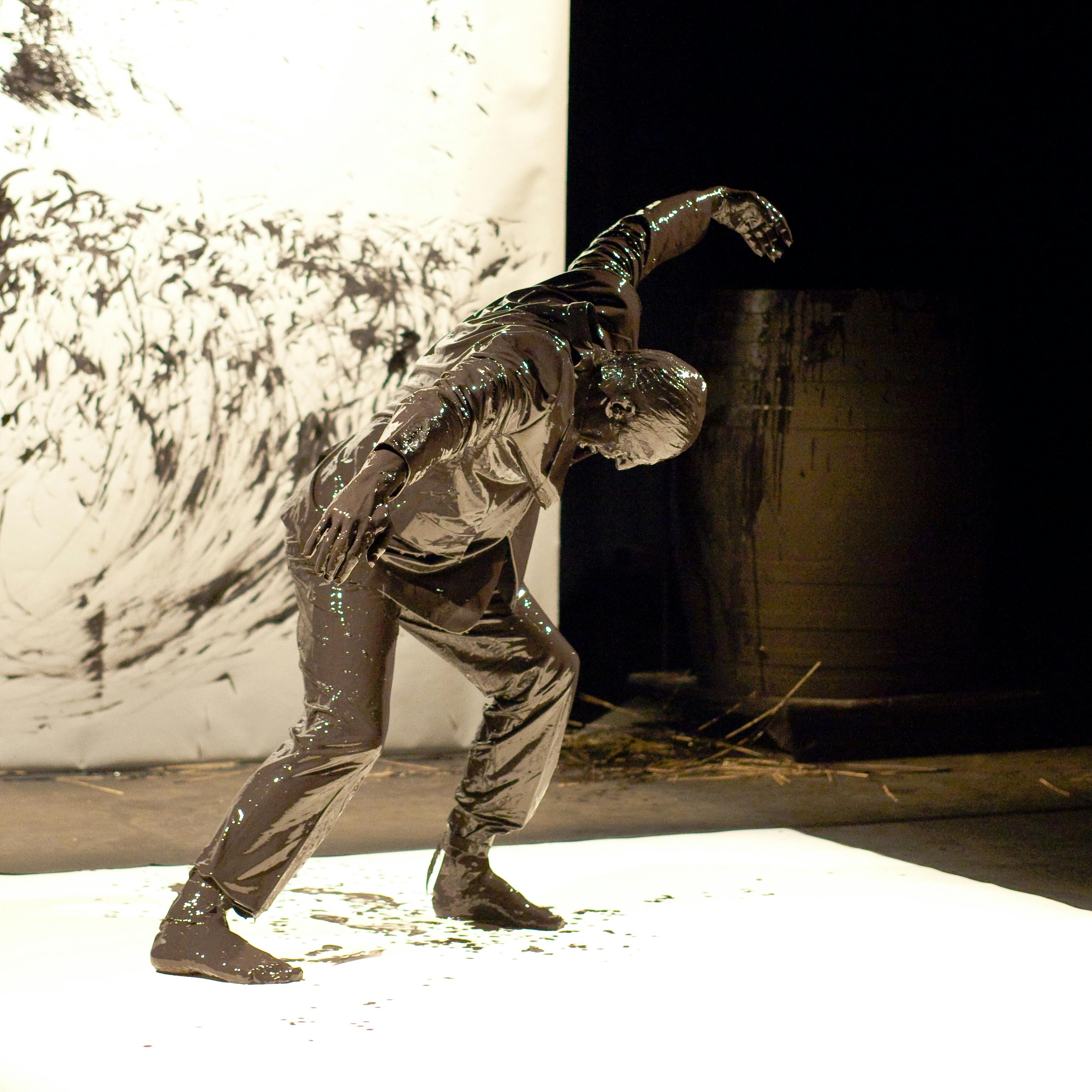 An artist in motion, completely covered in brown paint. On the ground and in the background, white sheets.