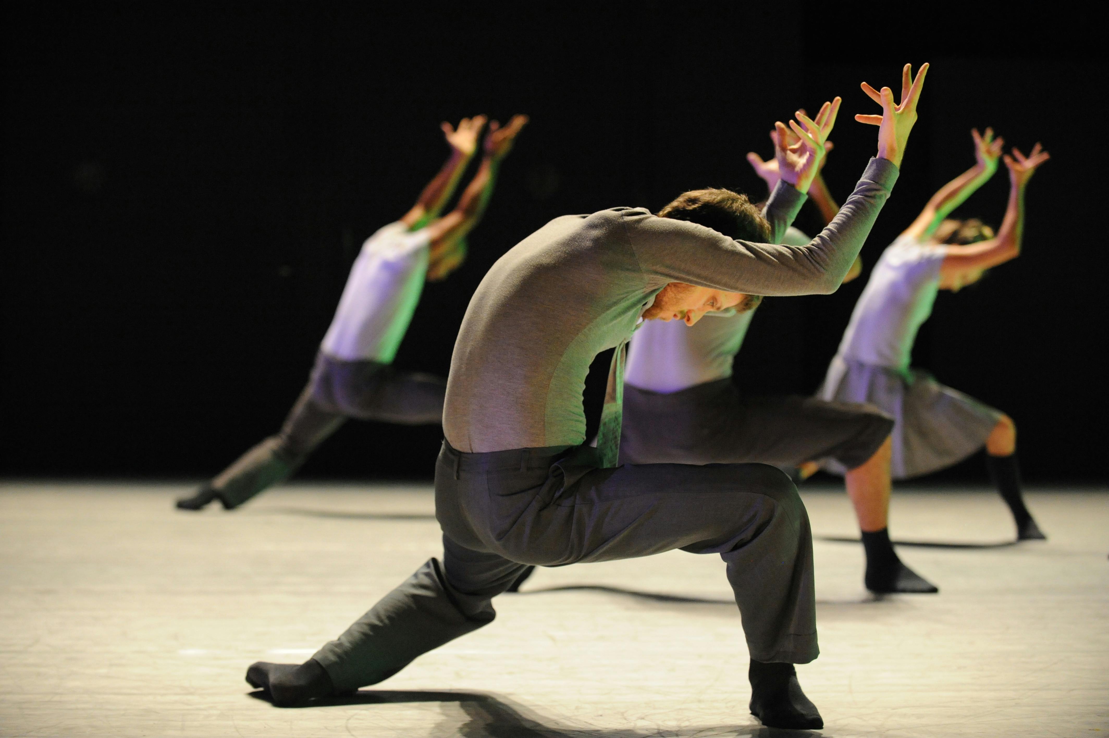 A side photo of four artists. They are bent forward with one knee almost touching the floor, looking down, and their arms stretched upwards.