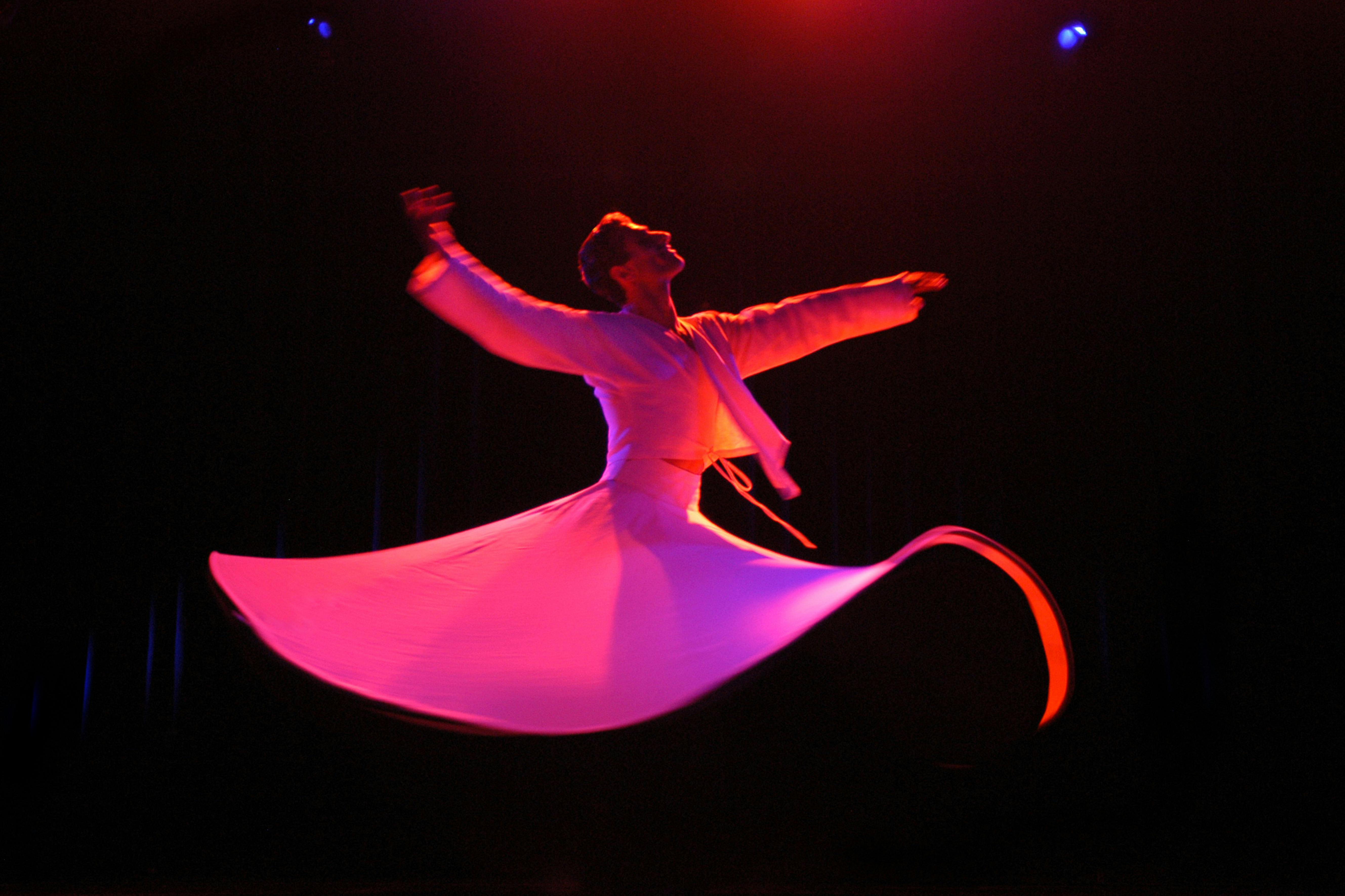 Foto di un ballerino. La luce è molto scura. Sta facendo una giravolta e la gonna che indossa è molto larga. Il vestito è illuminato con luce rosa.
