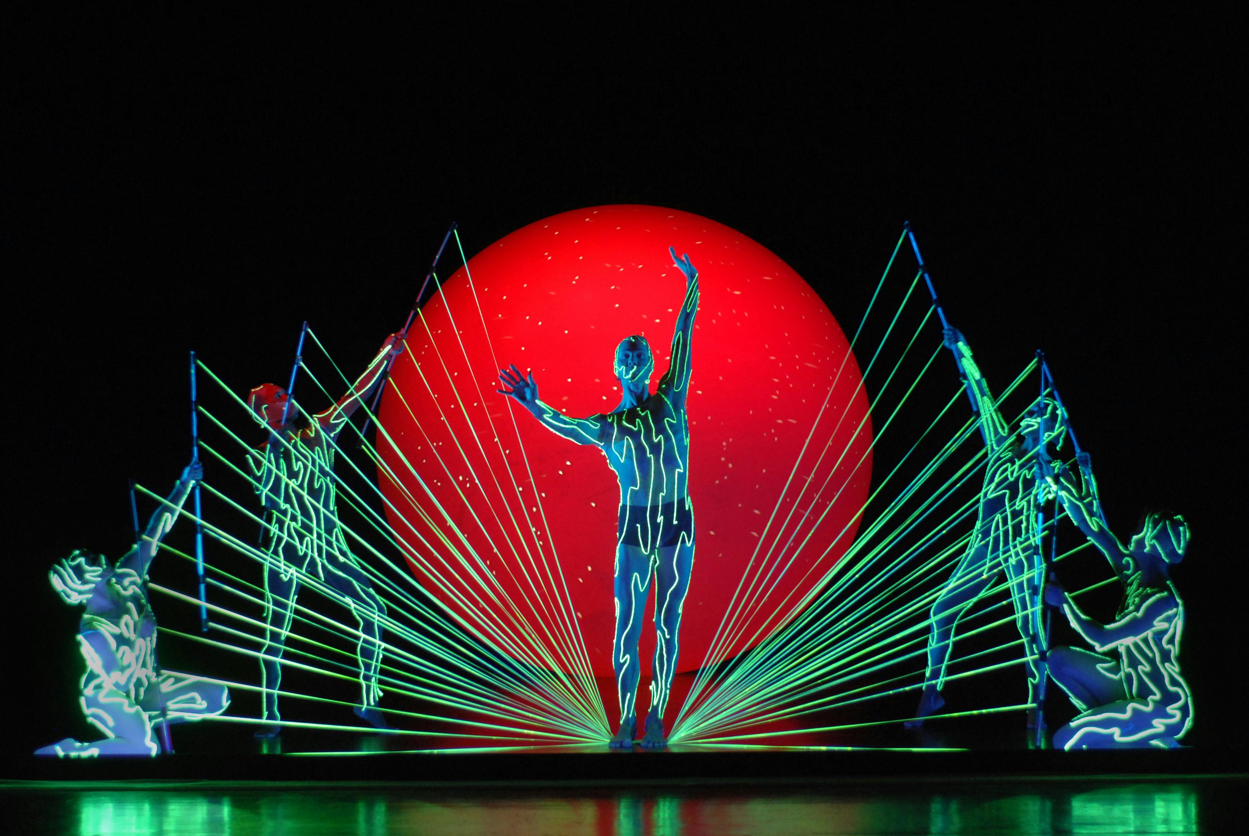 Five artists are on stage, wearing suits that glow in the dark. One stands in the center, while the others are positioned at the sides, pulling luminous ropes toward the one in the middle.