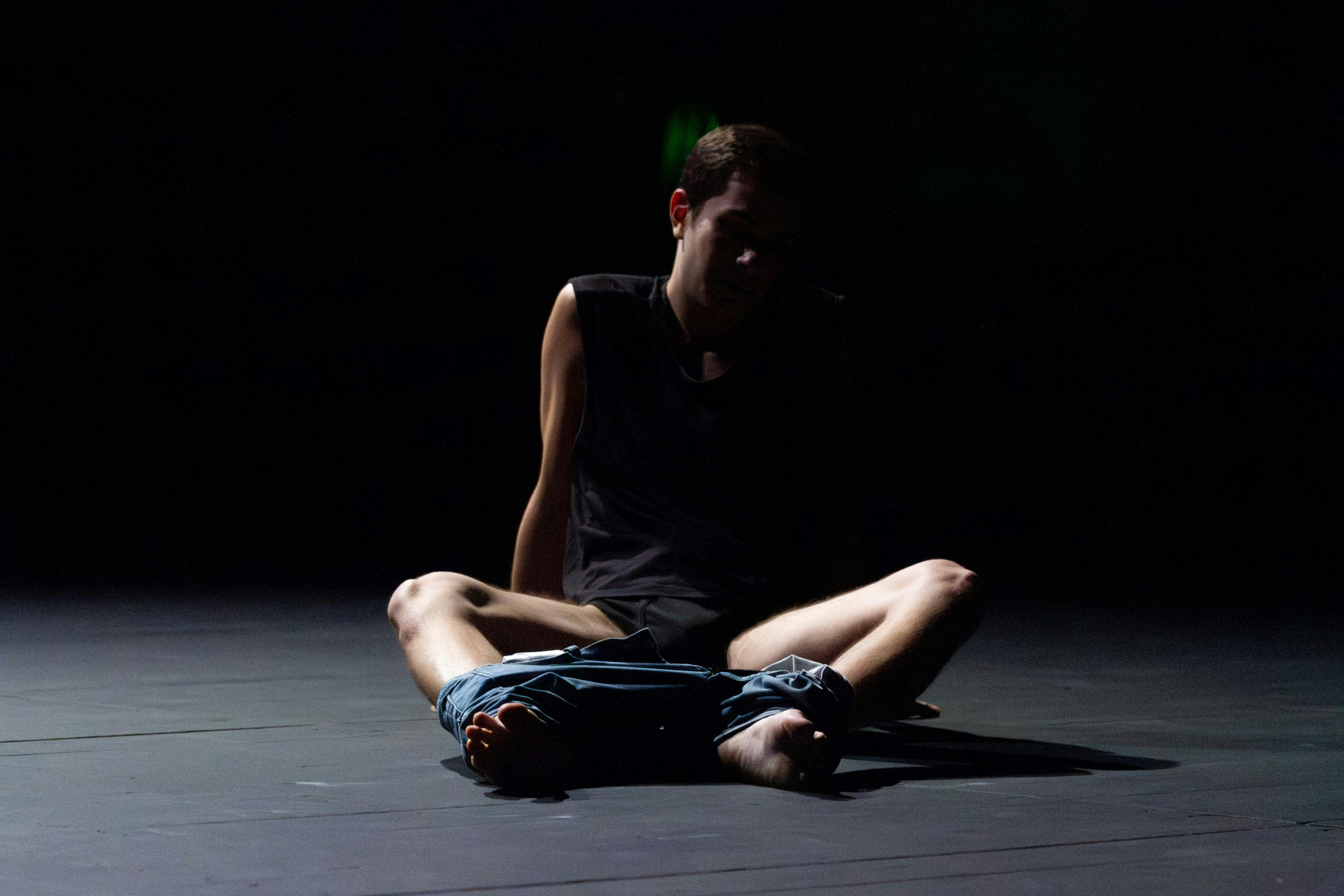Performer sitting on the floor in the half-light, legs slightly apart; wearing a black shirt and pants pulled down to ankle level.