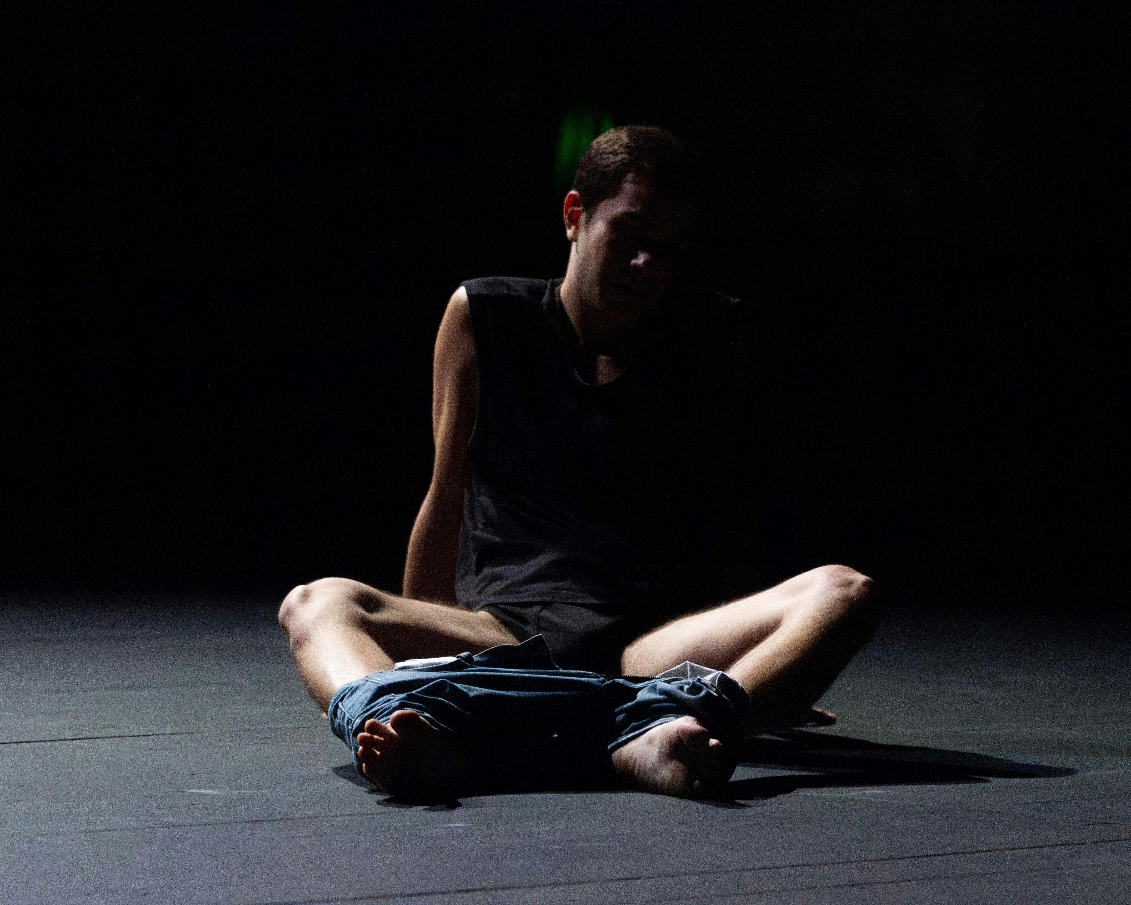 Performer sitting on the floor in the half-light, legs slightly apart; wearing a black shirt and pants pulled down to ankle level.