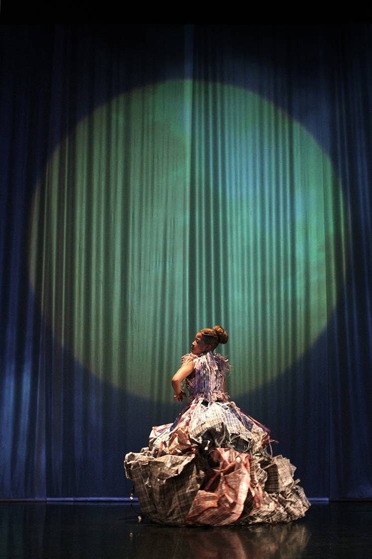 Sul palco una ballerina di spalle. Indossa un abito molto ampio. Sul sipario un grande faro giallo