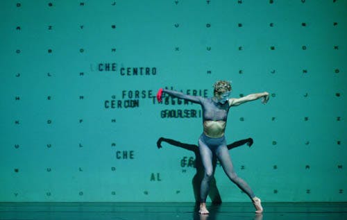 Una ballerina è in posa con le braccia tese e larghe e la gamba sinistra piegata. Sullo sfondo un fondale verde con delle scritte.