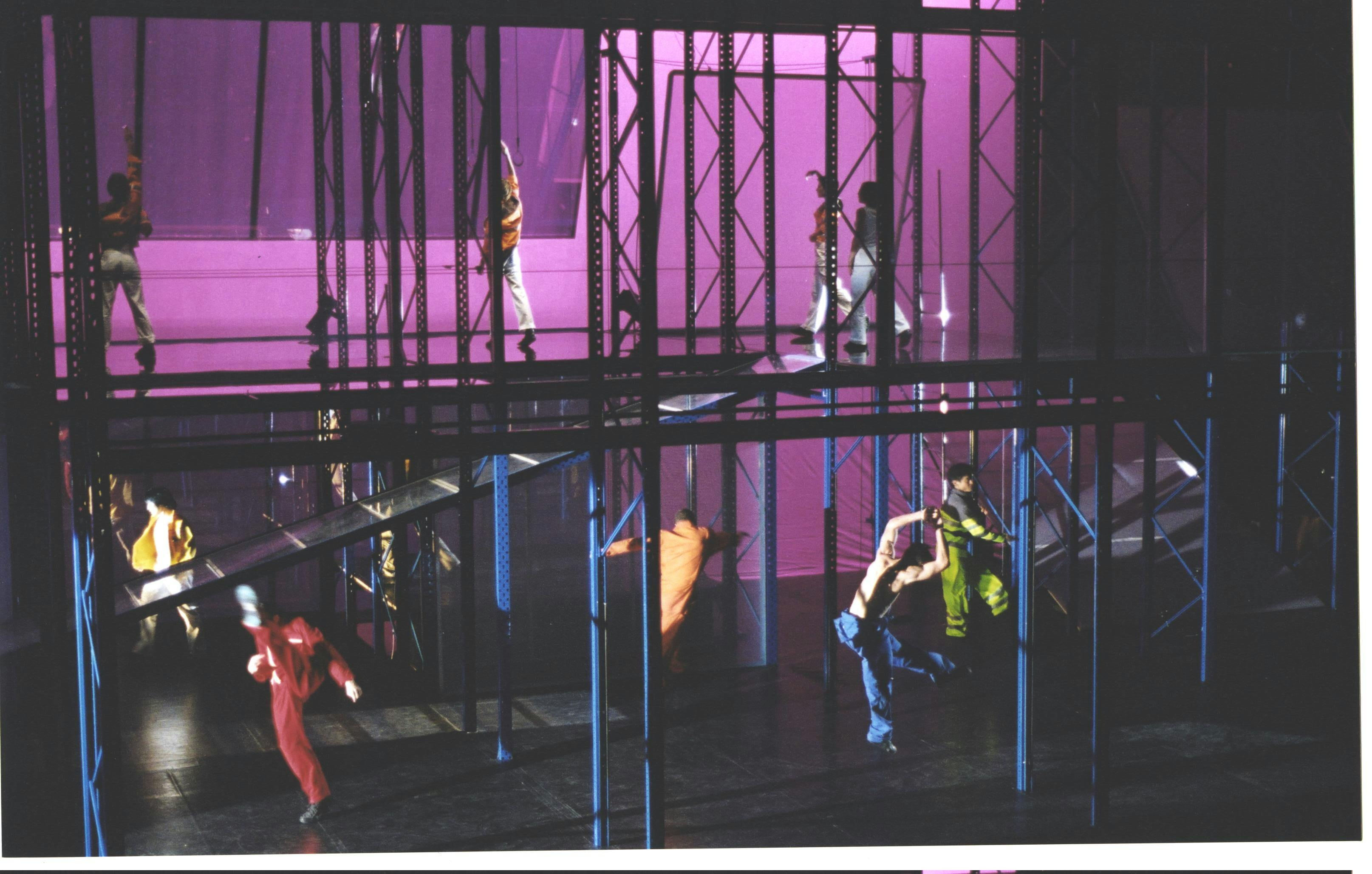 About ten artists dance amidst a two-story scaffold constructed with tubes.