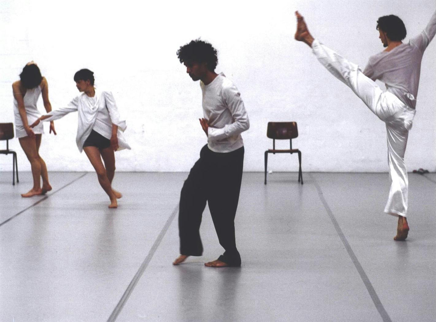 Quattro ballerini in sala, in pose differenti. Una a sinistra è di spalle e guarda in alto, un'altra corre. Quello al centro sembra camminare, quello a destra tende la gamba in alto.