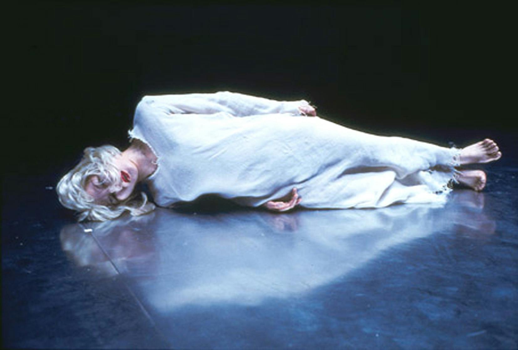 Una ballerina è sdraiata a terra, di lato. Indossa un abito bianco, ha capelli biondi e rossetto rosso.