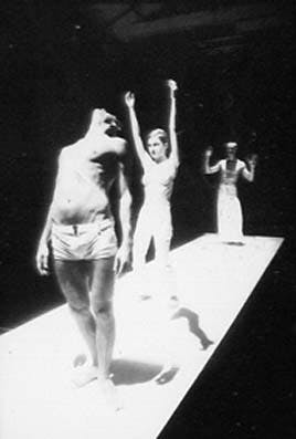 Three artists on a white catwalk. The first one is in underwear, looking up. The second one has her arms stretched upwards. The third one, at the back, raises his arms slightly.