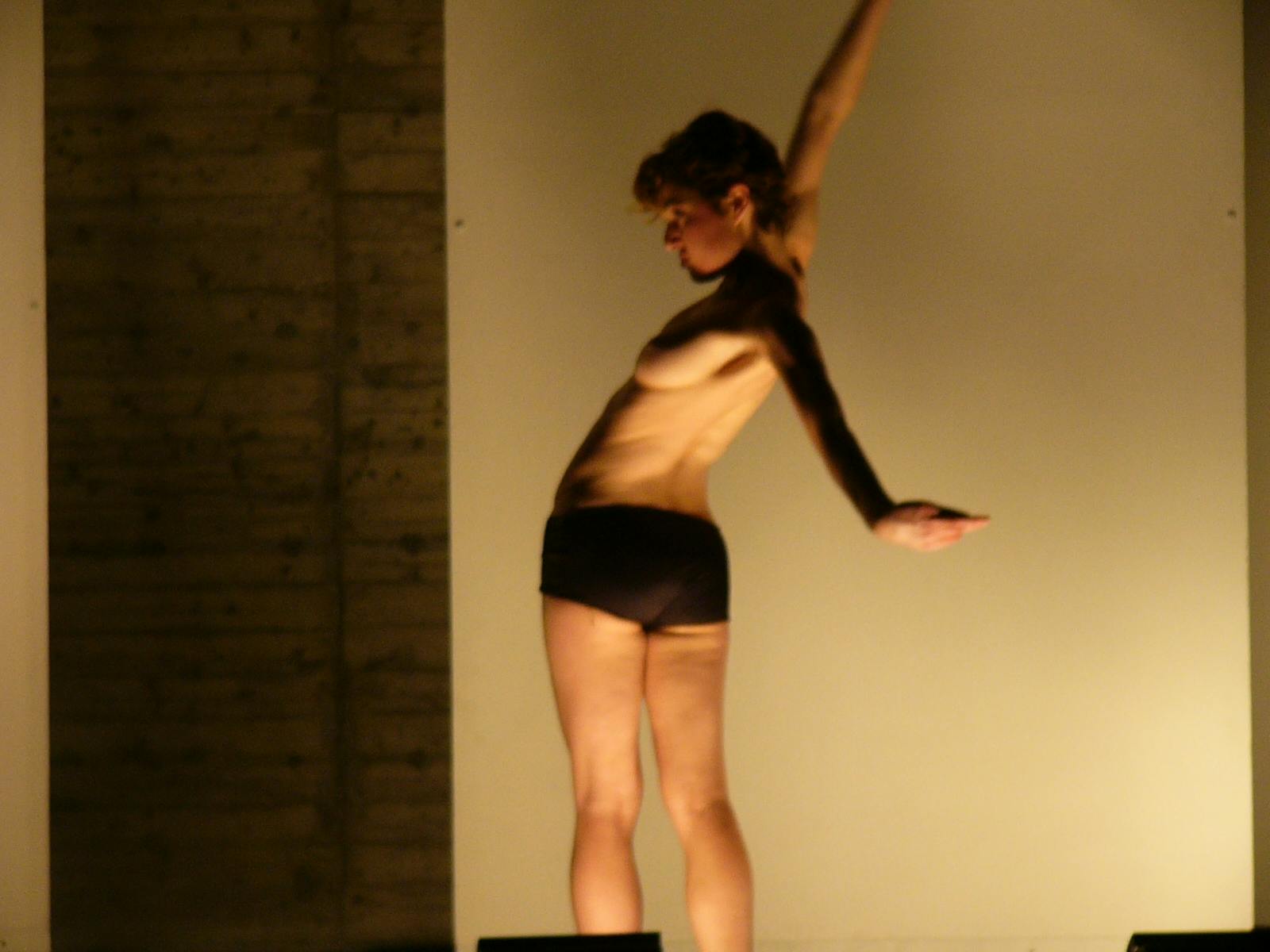 Una ballerina, con il petto nudo, di schiena guarda alla propria sinistra con il braccio destro teso in aria e il braccio sinistro teso all'indietro.
