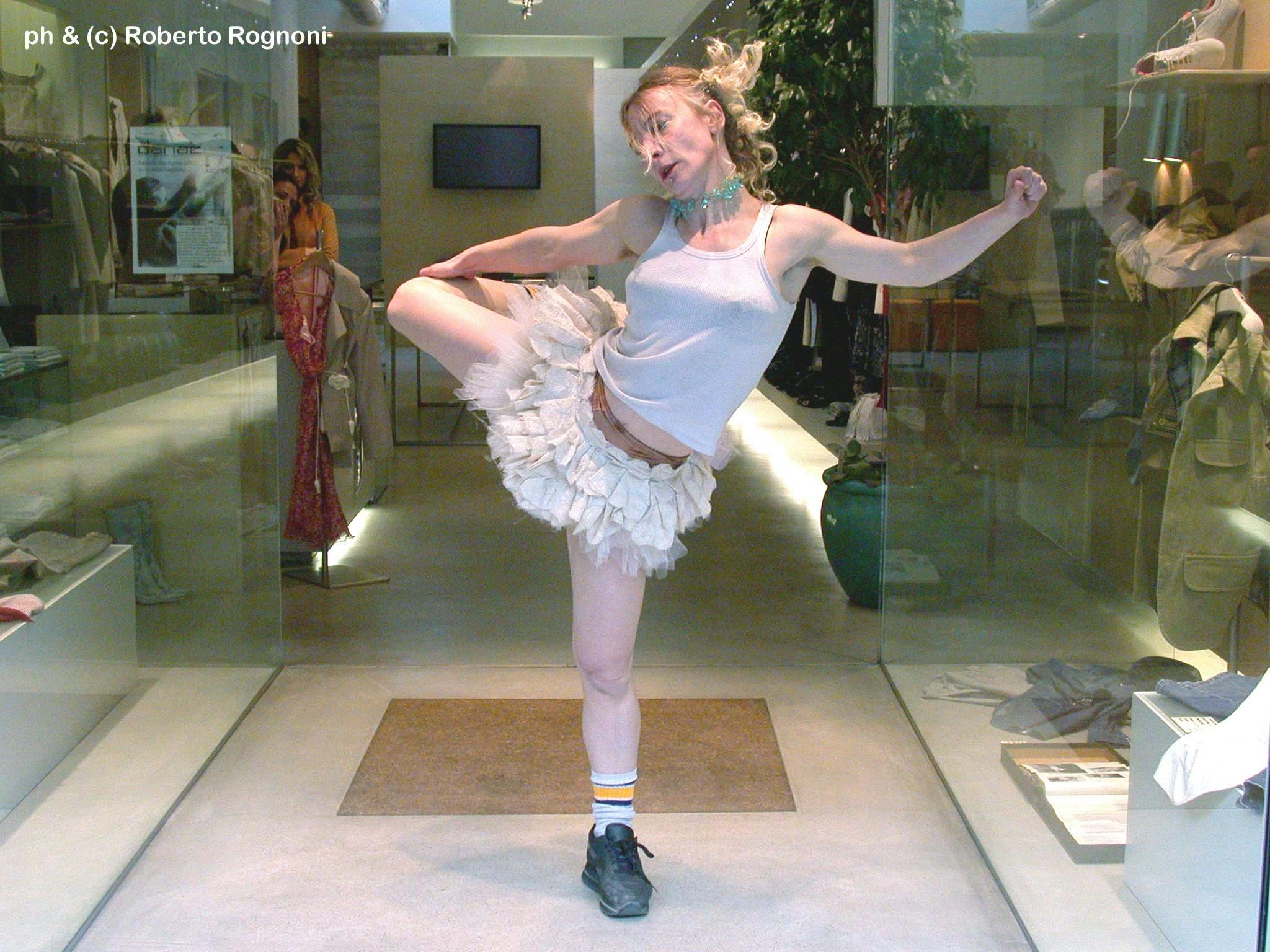 A ballerina, dressed in a white tutu and black sneakers, posing between two clothing store windows.