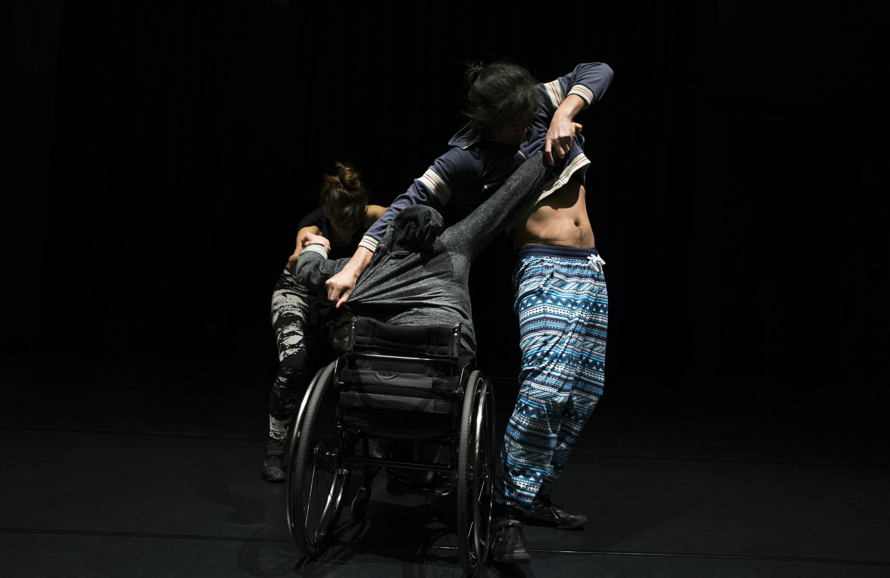 Three artists on stage. One is in a wheelchair, facing away. Another artist is in front of him, pulling him. Another is standing, pulling the arm of the first artist.