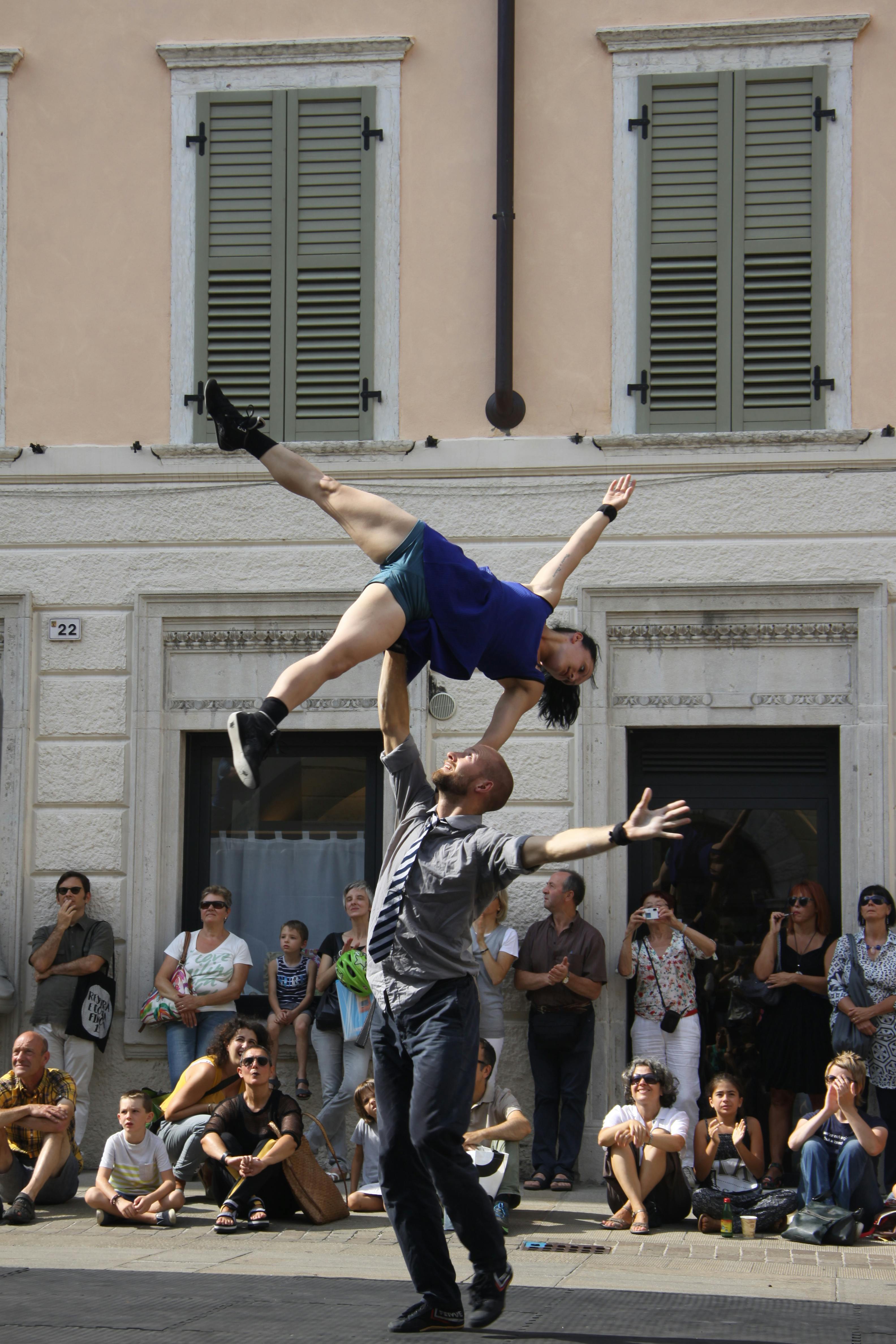 In the square in Rovereto, two artists. A woman is balancing on the outstretched arm of a standing man.