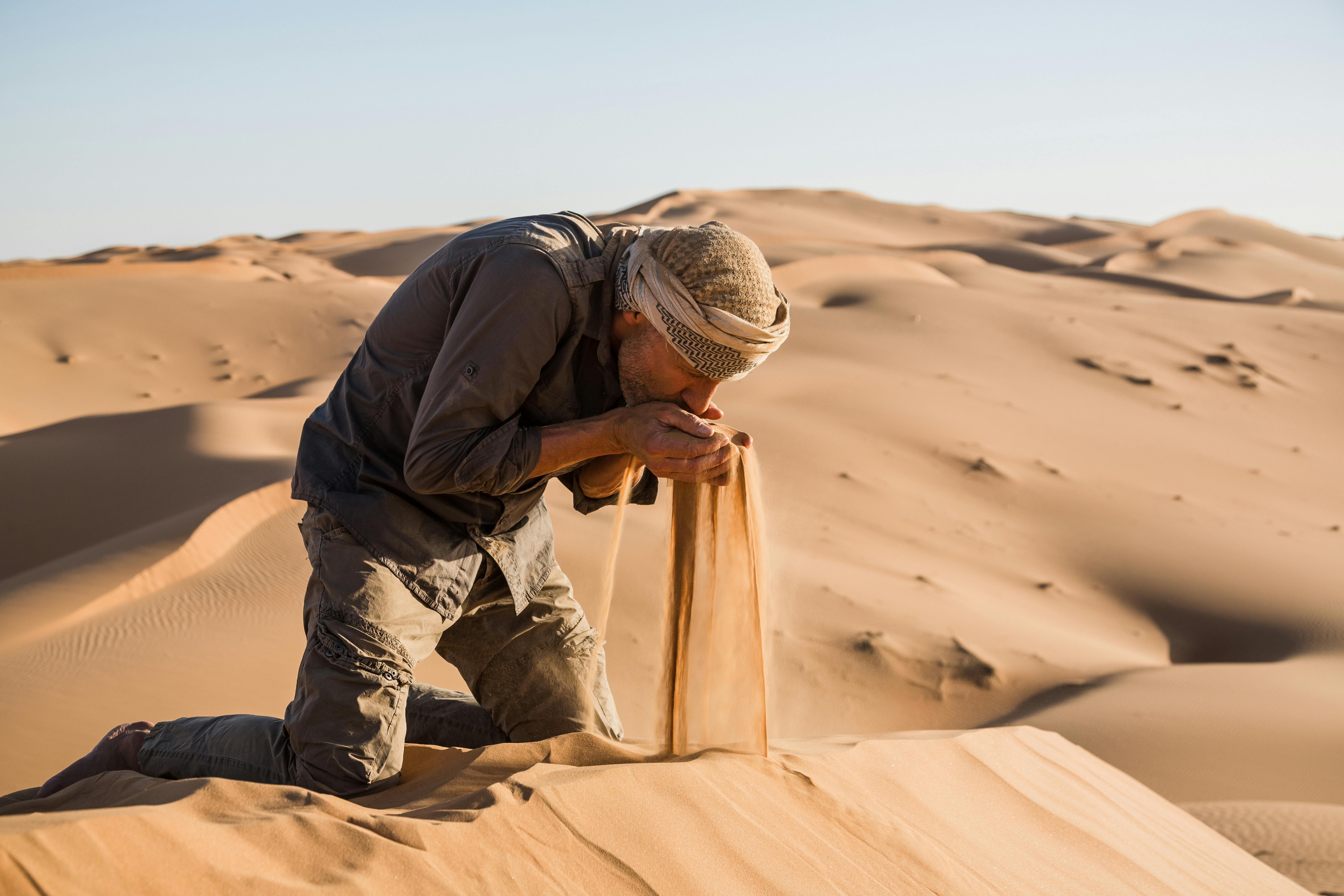 Photo of Max Calderan kneeling in the middle of the desert dunes, appearing to eat sand.
