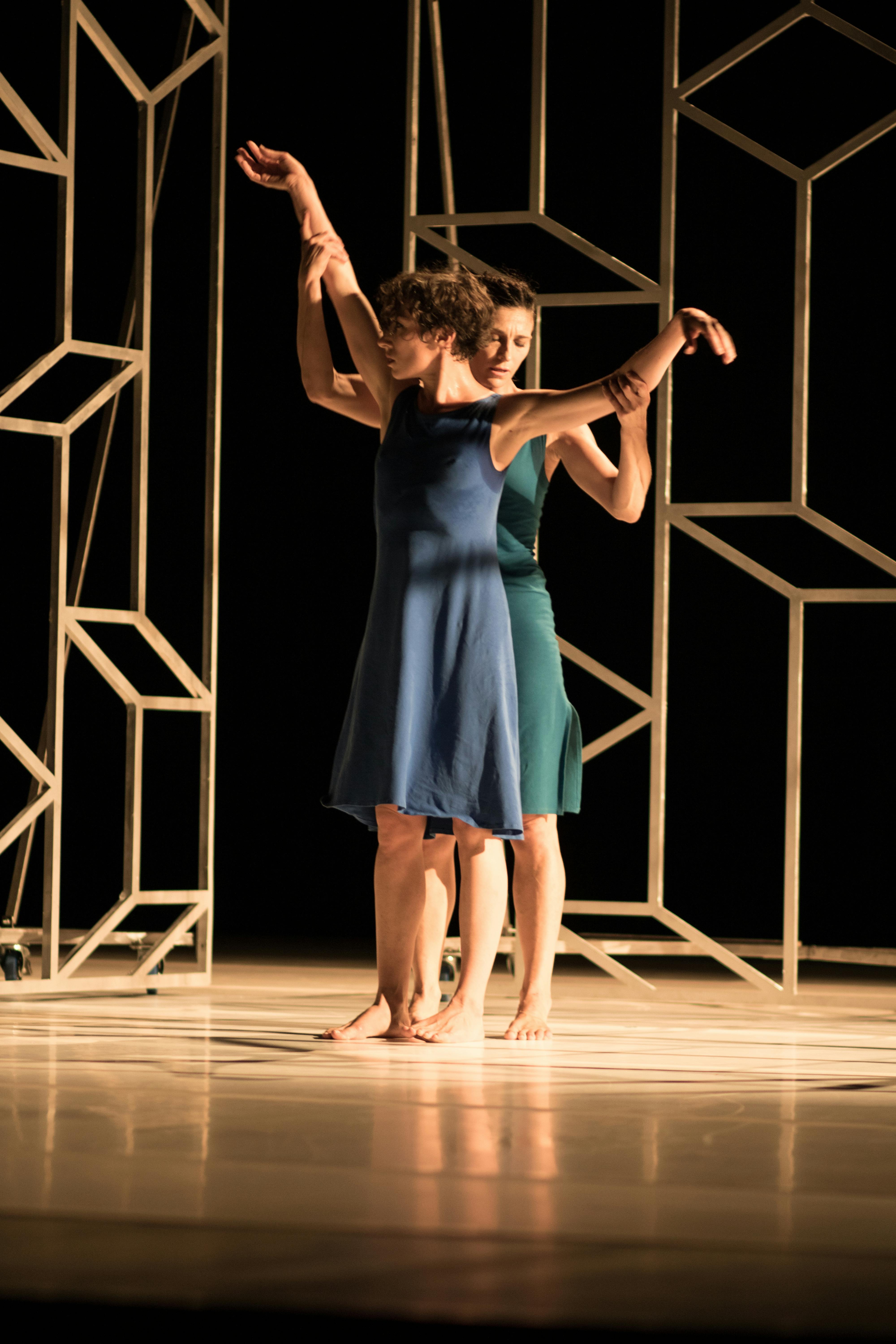 Two dancers are on stage, wearing knee-length blue dresses. They are positioned one behind the other, with the one in the back holding the arms of the dancer in front. Behind them, there are wooden structures.