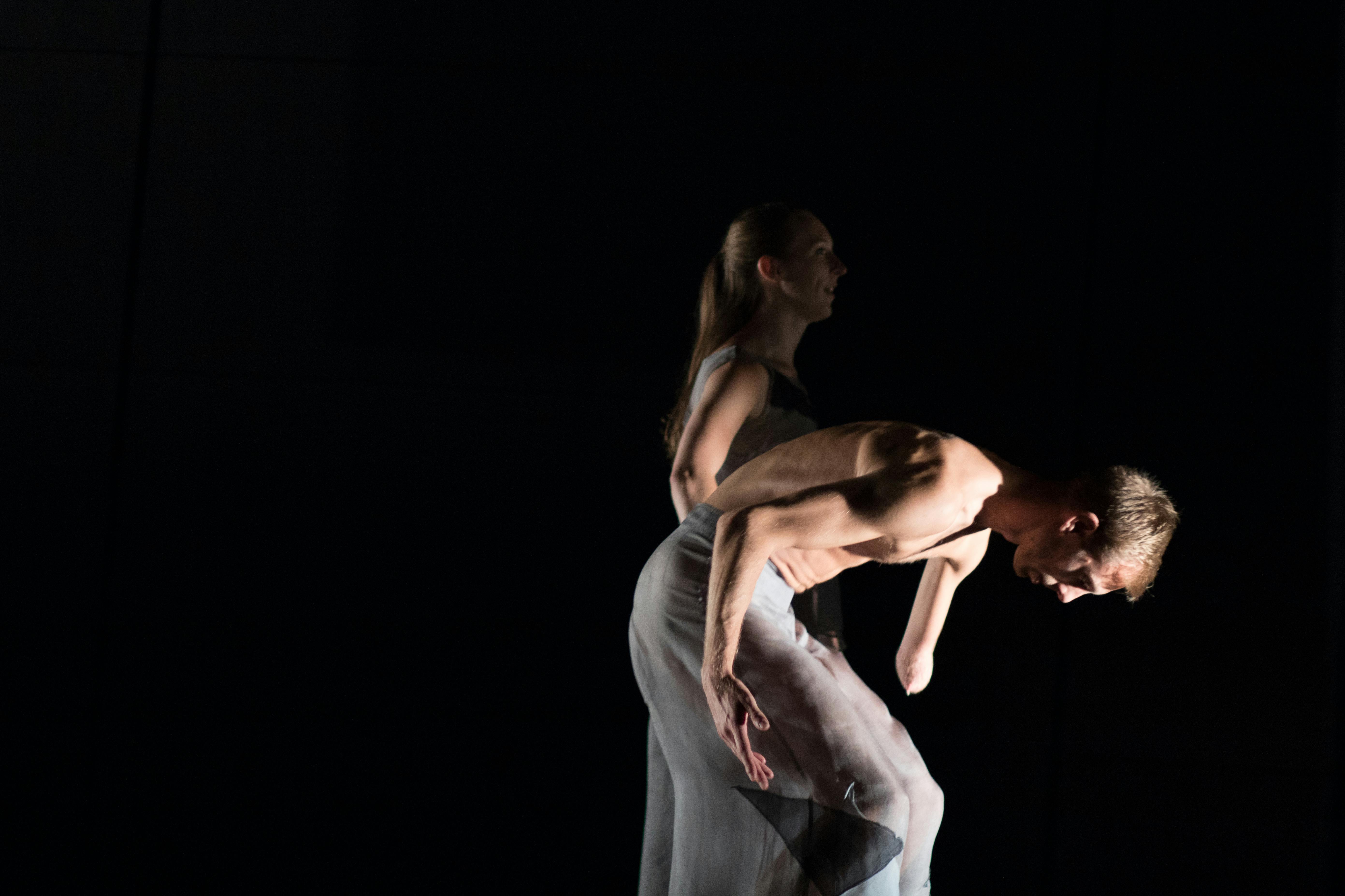 Profile picture of two artists. The one in front is wearing a tulle skirt, is bare-chested, and is leaning forward. They have only one arm. Next to them stands another artist.