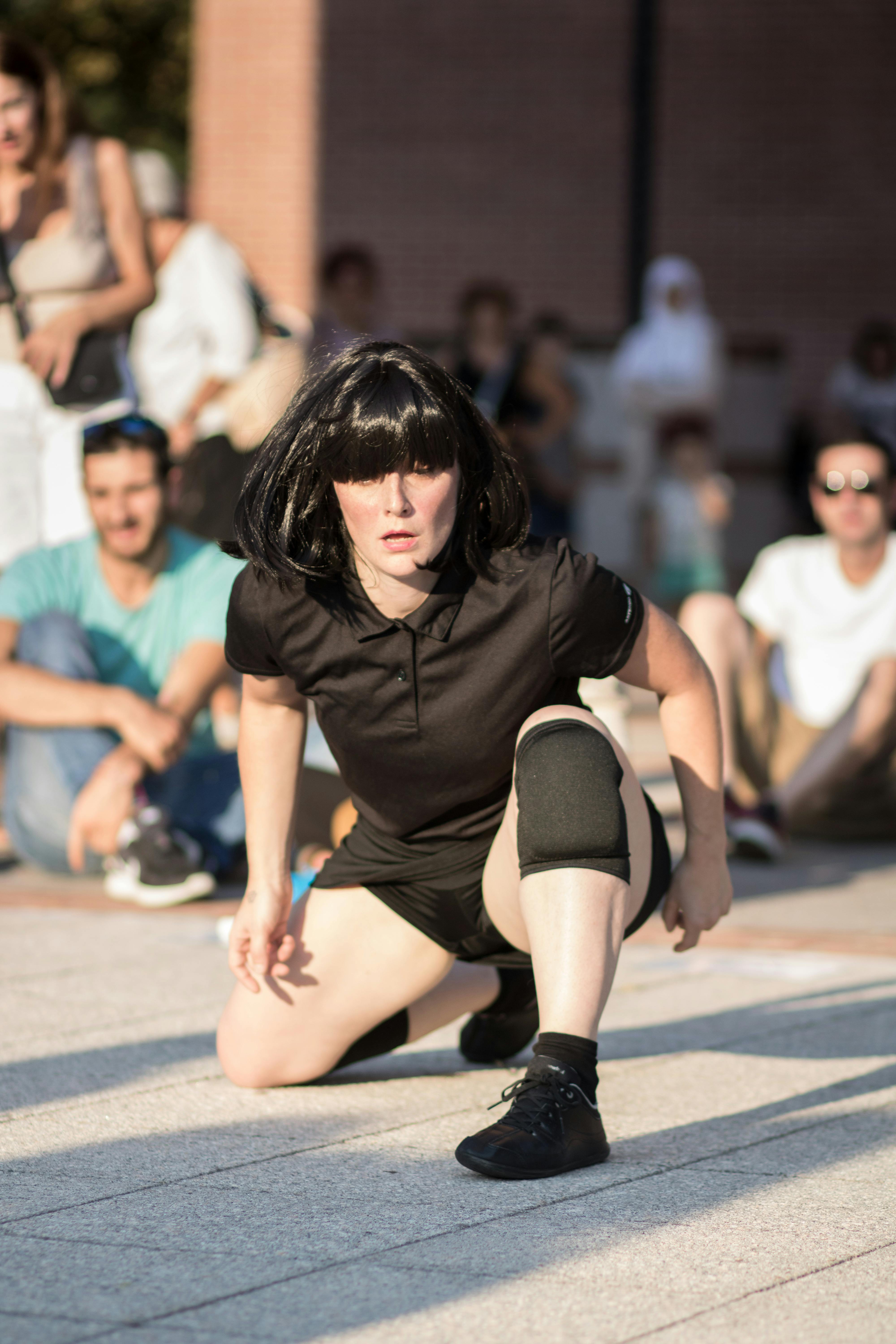 A dancer is in the square. She is wearing hair, a t-shirt, shorts, and black sneakers. She is kneeling on one knee and looking straight ahead. Behind her, there is an audience seated on the ground.