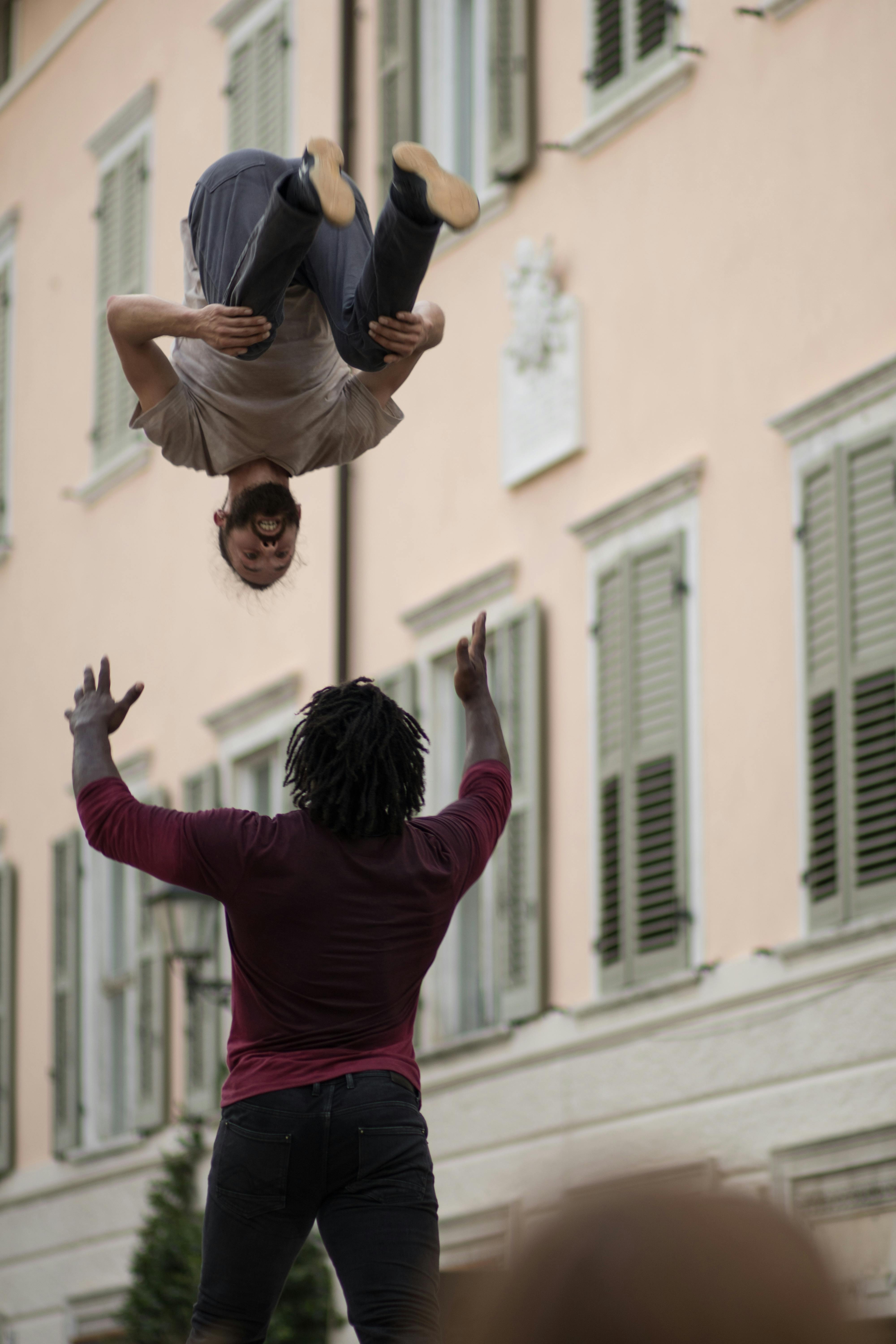 An artist is suspended upside down, holding their knees to their chest. Below them, another artist is ready to catch them.